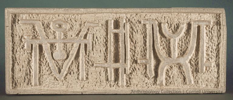 VOTIVE OF STILT-WALKERS | Photo by Ann Cady, 2024 | ©2026 Norman Daly Art — The artifact is comprised of a rectangular, textured stone slab featuring intricate carvings of abstract symbols and shapes, reminiscent of ancient writing or hieroglyphs, meticulously etched into its rough surface; the slab exhibits a border framing the artistic symbolism, and the overall tone is an earthy beige, creating a contrast between the raised, smooth carvings and the rough, indented background, suggesting a historical or archaeological significance, possibly meant to convey messages or stories from a bygone era.