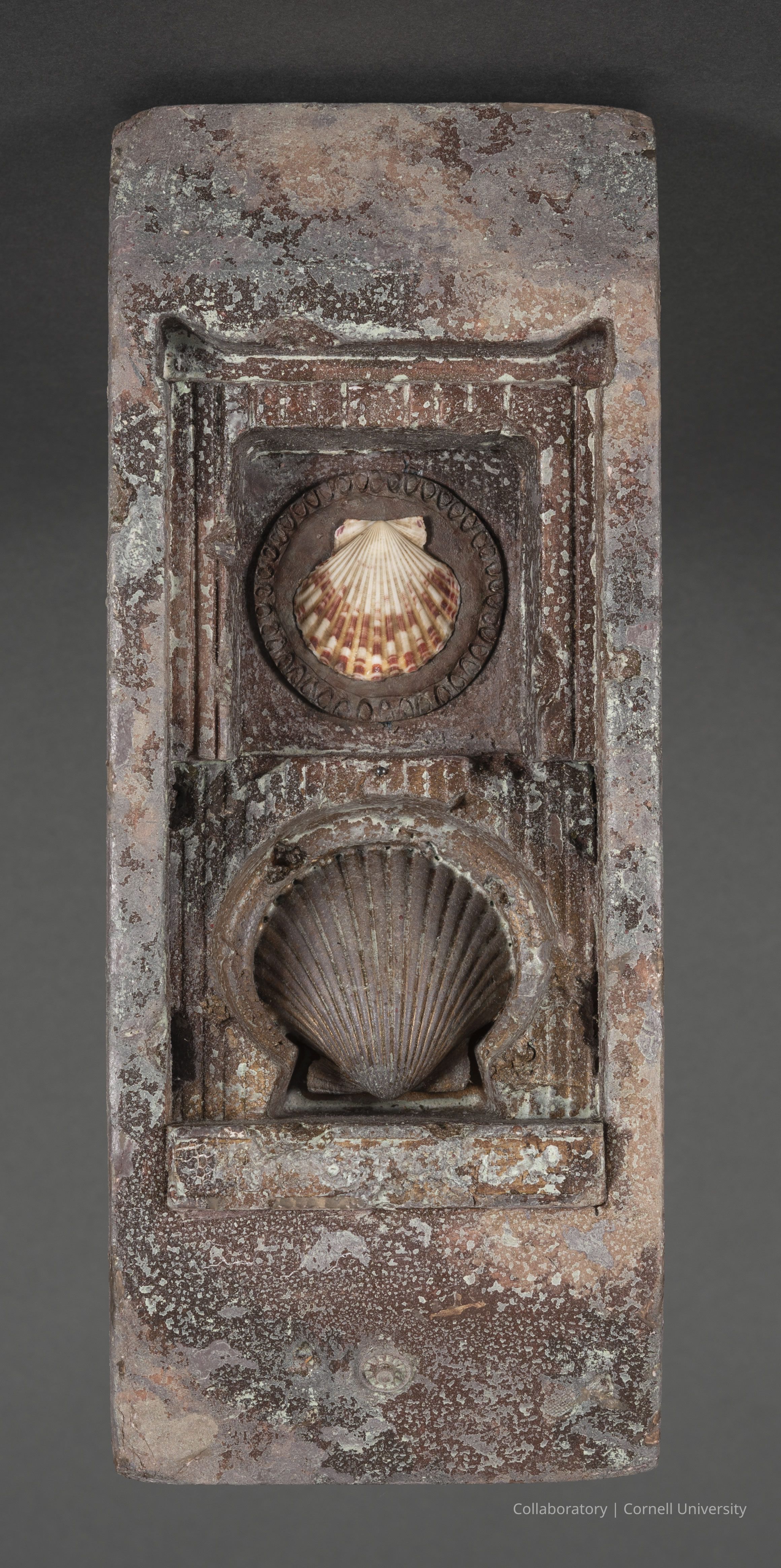 BODROCHNA (Combination Fertility Votive and Menstrual Chart) | circa 1966-69 | Photo by Ann Cady, 2024 | ©2025 Cornell University: The photo depicts an unusual artifact--a weathered stone slab featuring two carved clam shells within decorative frames. The upper shell is patterned and lighter--more pronounced--while the lower shell appears darker and blends with the stone. The overall texture of the slab is rough, with visible signs of erosion and aging, displaying a mix of brown, gray, and white hues. The intricate detailing around the shells holds the viewers attention.