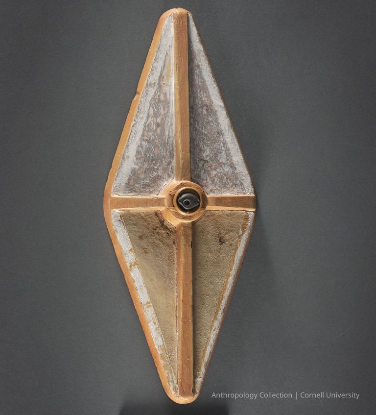 UNIDENTIFIED | circa 1966-71 | Photo by Ann Cady, 2024 | ©2026 Norman Daly Art — The image depicts an artifact with a diamond-like shape and a distinct convex form, featuring a symmetrical cross pattern in the center. The surface exhibits varying textures and colors, including earthy tones, with the edges outlined by wooden strips, creating a contrast against the darker background. The central area displays a circular element adding depth and complexity to the design, while subtle tones of white and gray present a weathered appearance, highlighting its possible historical or cultural significance. The craftsmanship indicates careful attention to both functional and aesthetic aspects.