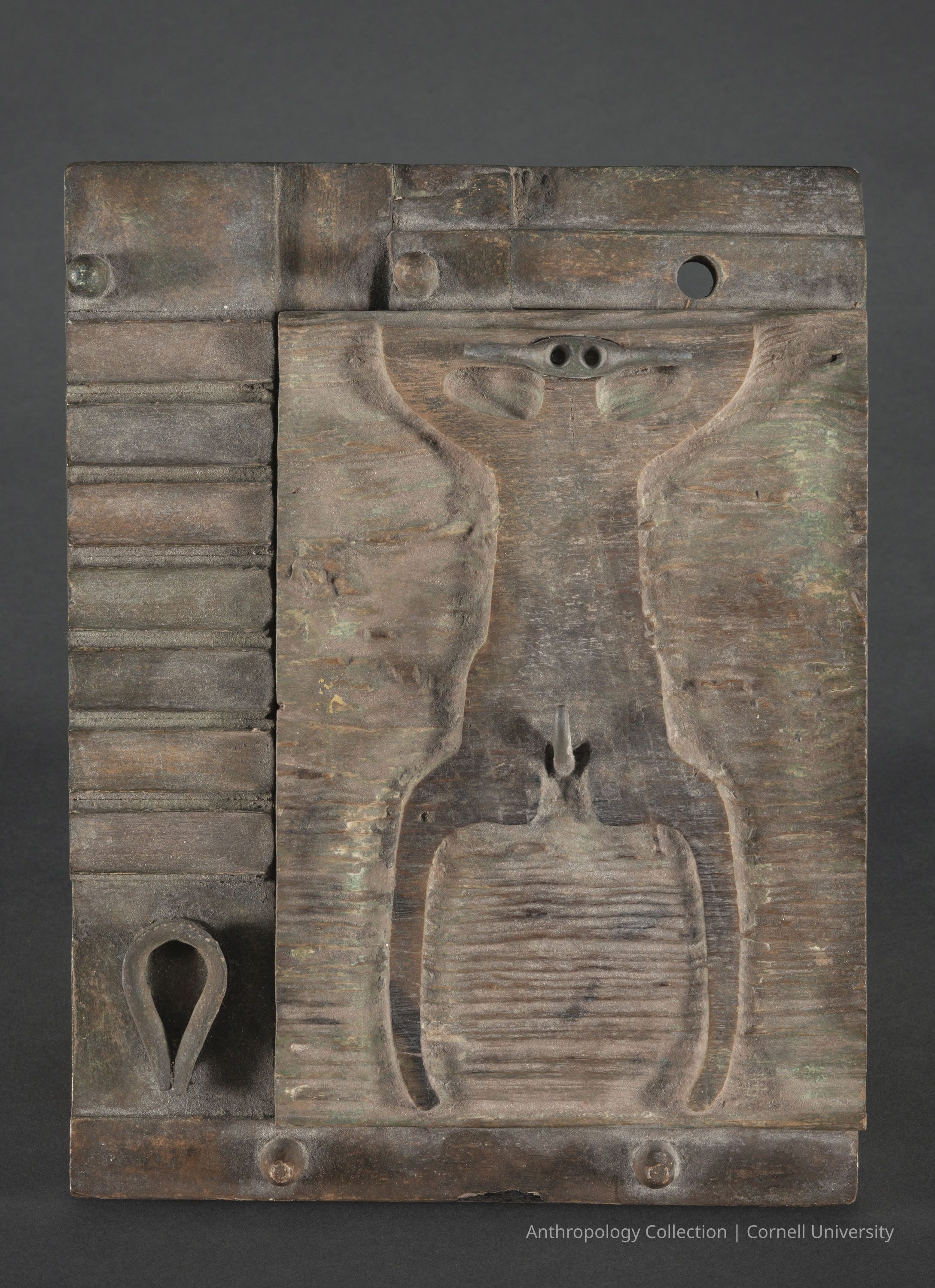 ARKPHOR OF THE SHRINE OF MALMUS (Temple Votive) | circa 1966-71 | Photo by Ann Cady, 2024 | ©2026 Norman Daly Art: A wooden plaque featuring a symmetrical design with a tribal influence, depicting what appears to be a stylized human figure. The artifact shows considerable age with a patinated surface, detailed with vertical ridges on one side and intriguing motifs, cut-outs and other features that create a mysterious, totemic appearance. The texture and tone suggest extensive use, giving this object a rich historical character possibly linked to cultural rituals or symbolic representations.