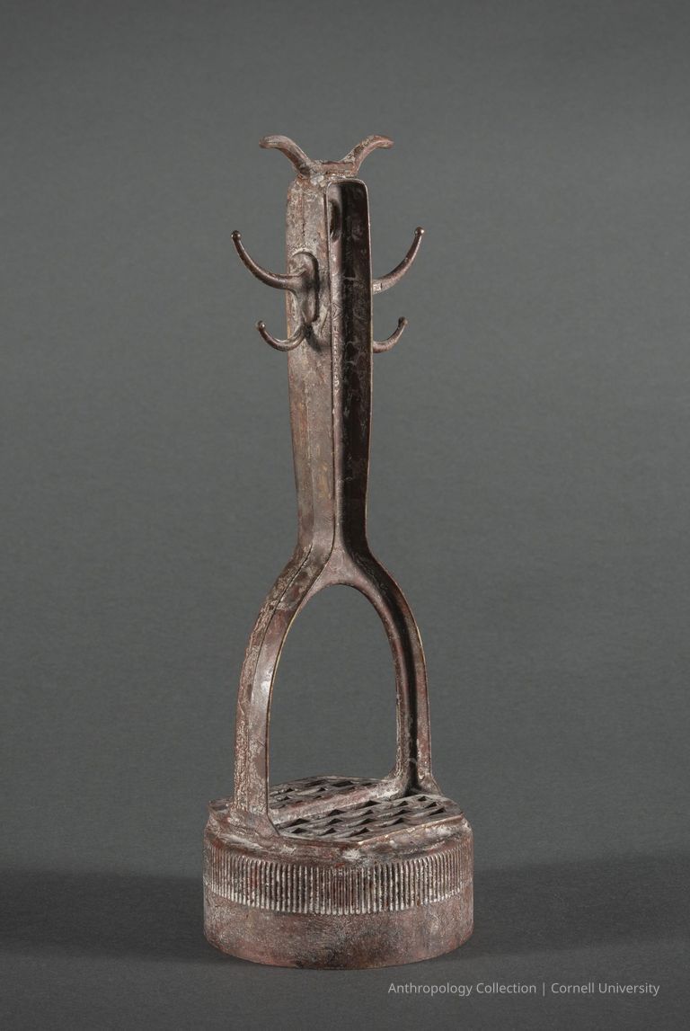 DWARF MONSTER | 1966-71 | Photo by Ann Cady, 2024 | ©2026 Cornell University — The image displays a rusted metal artifact representing the iconic ceremonial dwarf monster of Llhuroscian lore. It features a distinctive head with horns or antlers and two pair of arms. Its elongated body is seated on a rounded, grille-like base with ribbed detailing.