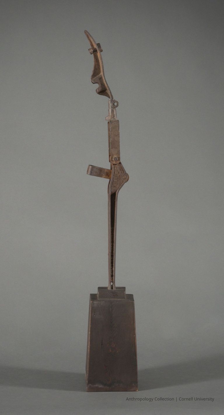 VOTIVE FIGURE | circa 1961-65 | Photo by Ann Cady, 2024 | ©2026 Norman Daly Art — A tall metal artifact, resembling an abstract humanoid figure with a protruding phallus.The figure's head, angled sharply upwards, is distinctive in its pointed, elongated shape, lending an aura of looking towards something distant or beyond.  It stands erect on a wooden base that shows signs of aging and water damage.
