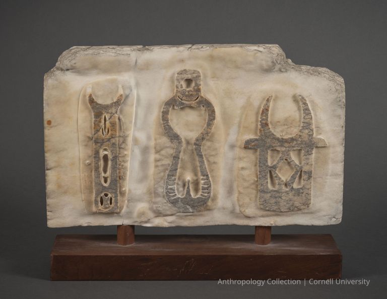 STELE WITH STILT-WALKER AND STILTS | circa 1961-65 | Photo by Ann Cady, 2024 | ©2026 Norman Daly Art — The image shows an ancient, weathered marble bas-relief featuring three vertical,  figures, each enclosed in an arched niche and intricately carved with symbols and geometric patterns, mounted on a simple dark wooden stand.