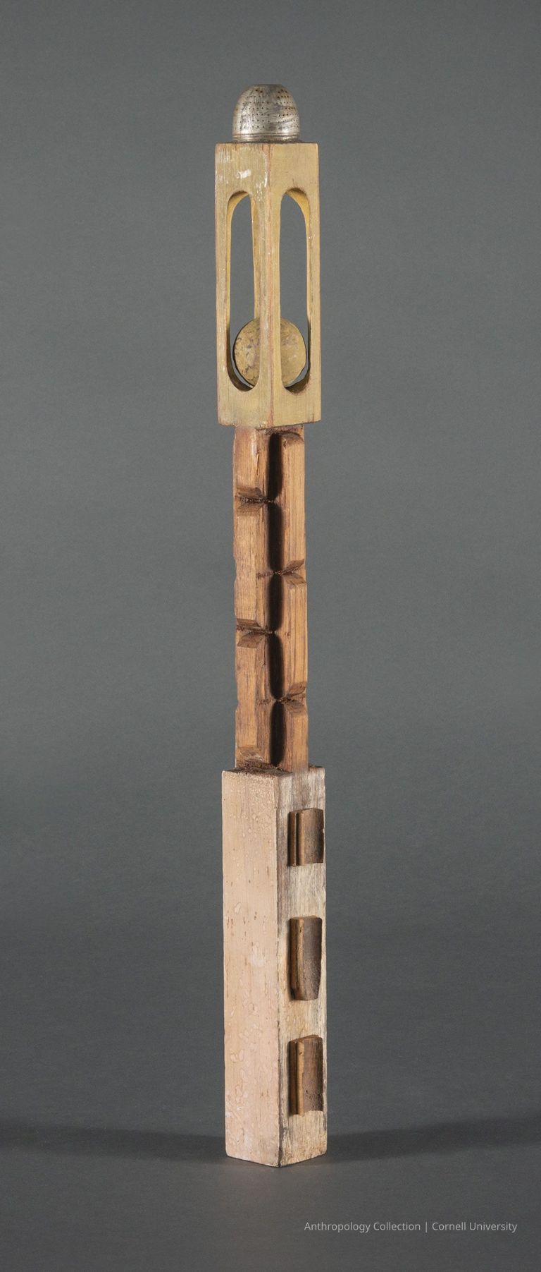 NOISEMAKER | 1966-71 | Photo by Ann Cady, 2024 | ©2026 Norman Daly Art — A long slender vertical wooden object with segments of rectangular cutouts. It features a dome-like metal piece at its apex, topping  an enclosure which holds a wooden ball appearing to be movable.