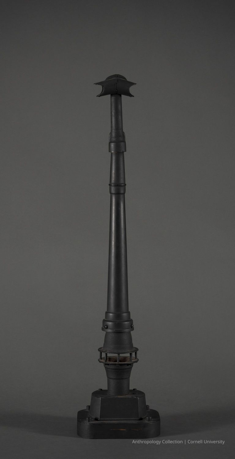 VOTIVE | 1965 | Photo by Ann Cady, 2024 | ©2026 Norman Daly Art — The image presents a tall, slender, dark-colored metal form resembling a stylized tower, topped with a curved, helmet-like structure. Each section of the form has distinct layers and textures, with a solid black base that grounds the piece against a grey backdrop.