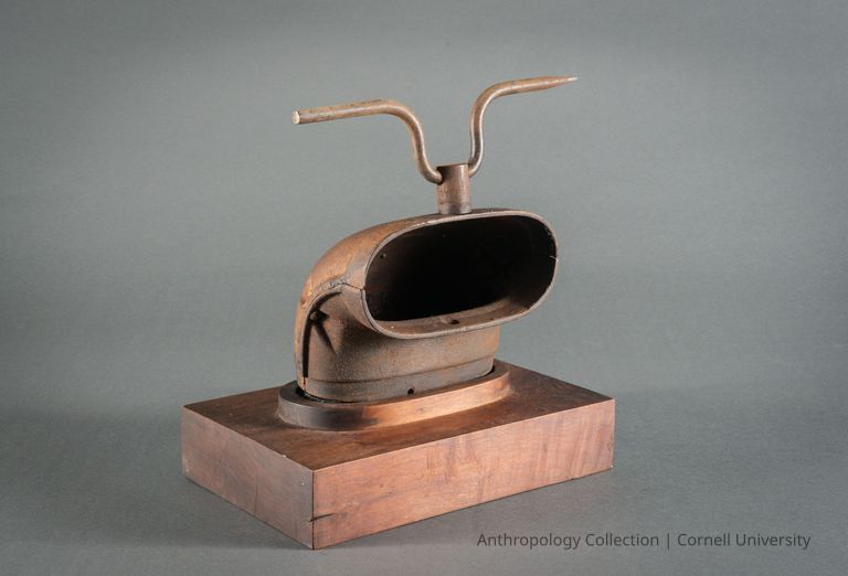 VOTIVE FIGURE | circa 1965 | Photo by Ann Cady, 2024 | ©2026 Norman Daly Art — The image shows a votive resembling a rusted metallic helmet with an open visor mounted on a wooden base; attached to the top is a bent metal rod that forms horns or antennae, portraying an abstract and intriguing artifact.