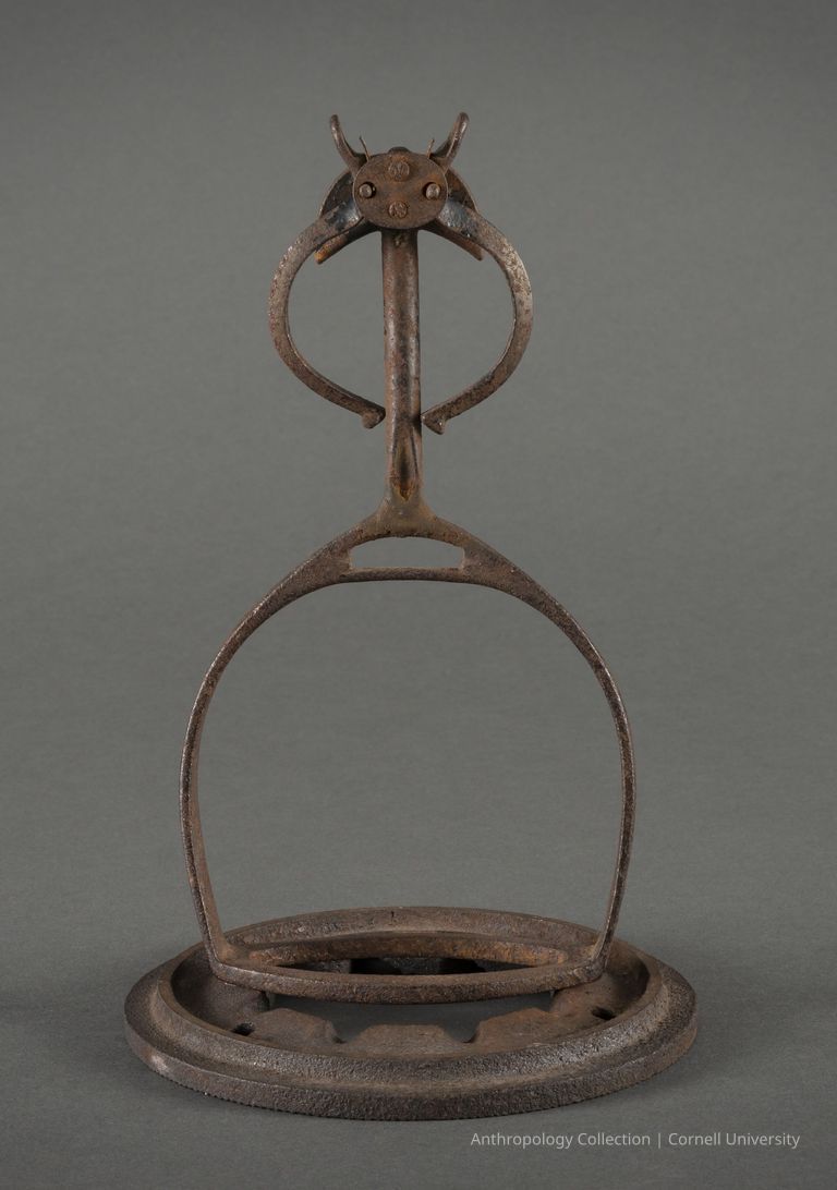 DWARF MONSTER RIDING A WHEEL | circa1966-71 | Photo by Ann Cady, 2024 | ©2026 Norman Daly Art — An rusted and weathered metal artifact with a circular base featuring a humanoid figure with bowed 'legs', and 'arms' akimbo and a 'face' with bulging eyes and horns.