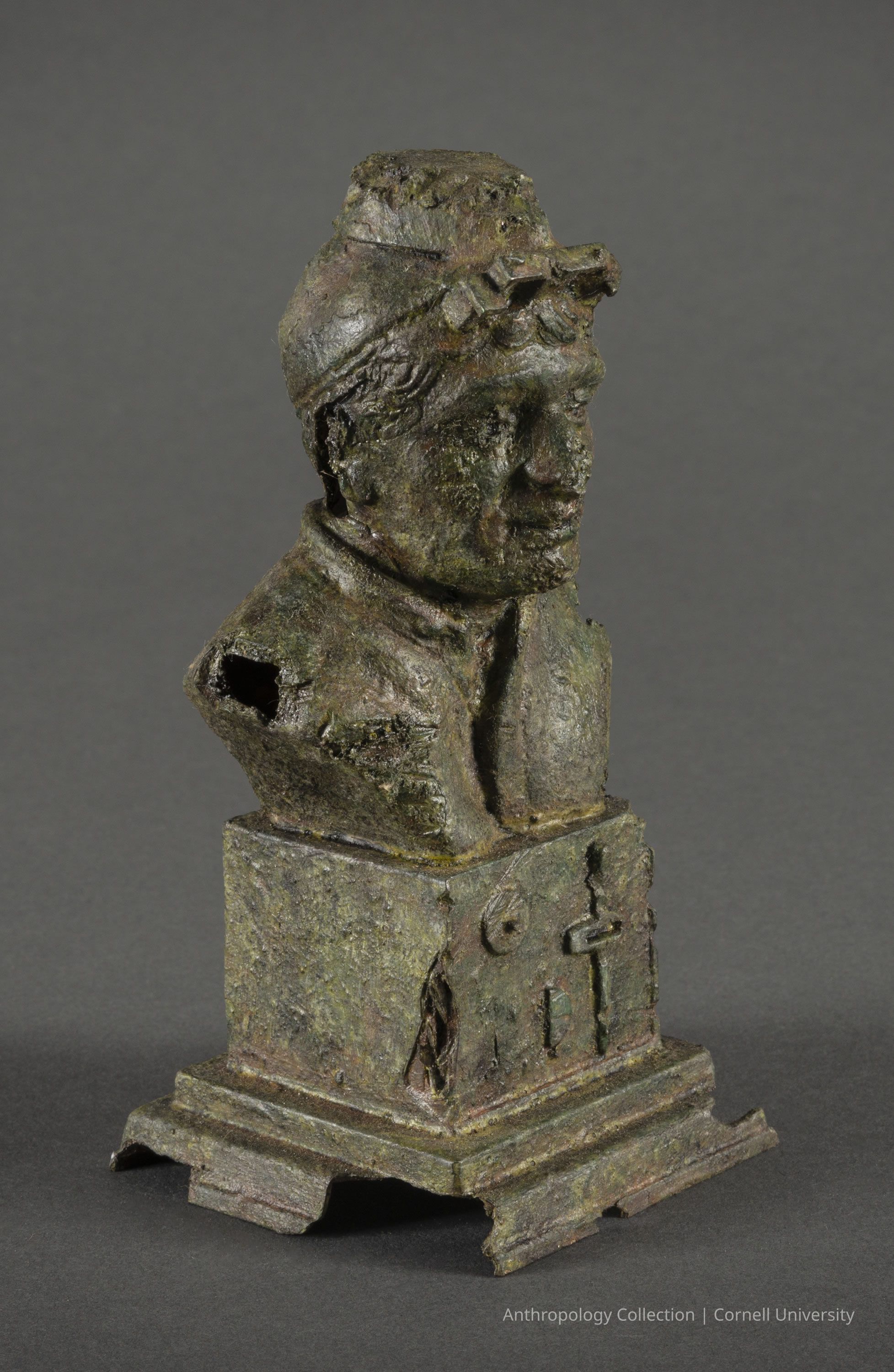 BUST OF DIRECTOR-REFEREE WITH THOUGHT TRANSMITTER | circa 1966-71 | Photo by Ann Cady, 2024 | ©2026 Norman Daly Art: An ancient, weathered bronze bust of a male Llhuroscian athlete, set upon a rectangular base with intricate carvings that suggest historical significance, symbolizing an era or civilization long past, evoking a sense of mystery and antiquity; the oxidized surface displays textures wrought by time.