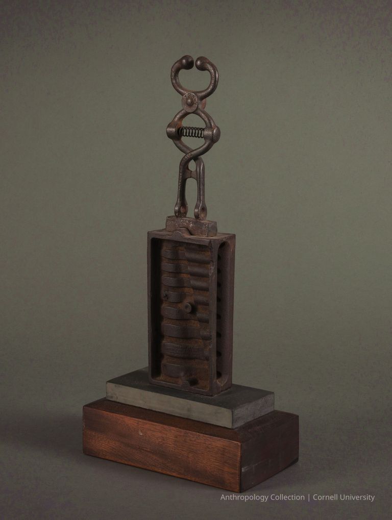 VOTIVE | circa 1961-65 | Photo by Ann Cady, 2024 | ©2026 Norman Daly Art — A metal artifact comprised of a rectangular section with vertically stacked, rounded segments, topped by a stylized humanoid-like metal figure with a spring-like torso and spherical elements at the top, possibly representing a head. The artifact is displayed on a two-tiered wood and slate base.