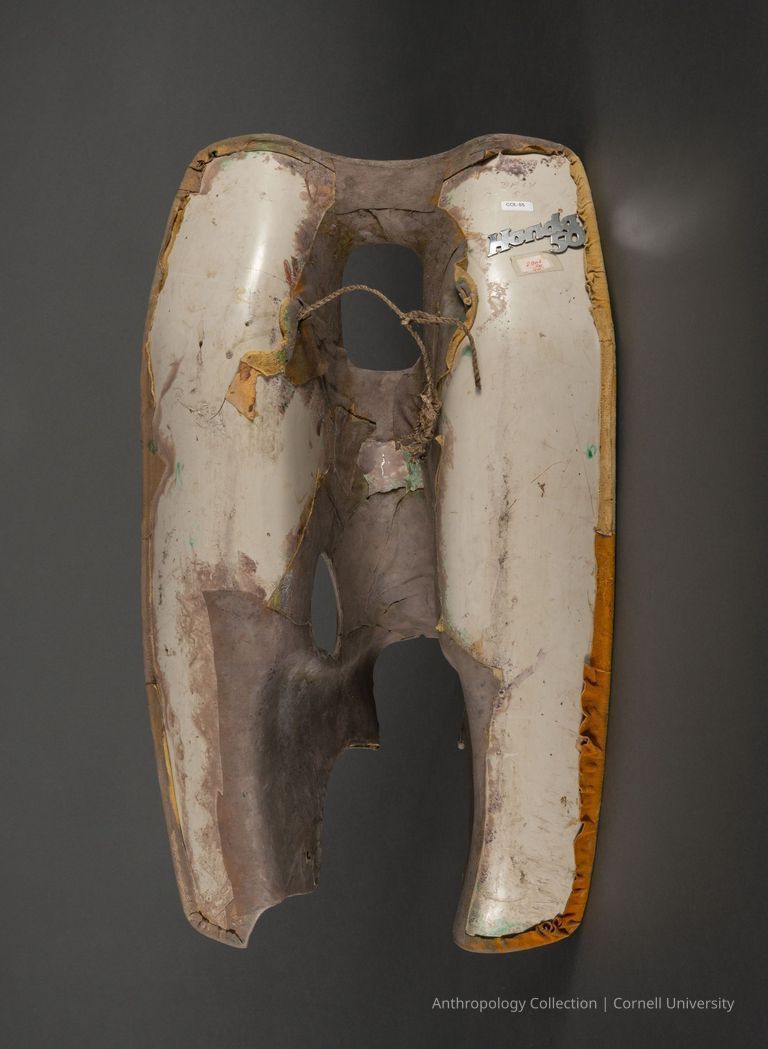 ELEPHANT MASK verso — The image depicts the front of an old, weathered motor scooter gas tank, covered with spots of paint and a frayed material, showing significant signs of wear and tear;  the piece is hanging against a plain, dark background, and there's a Honda emblem affixed to the upper right.