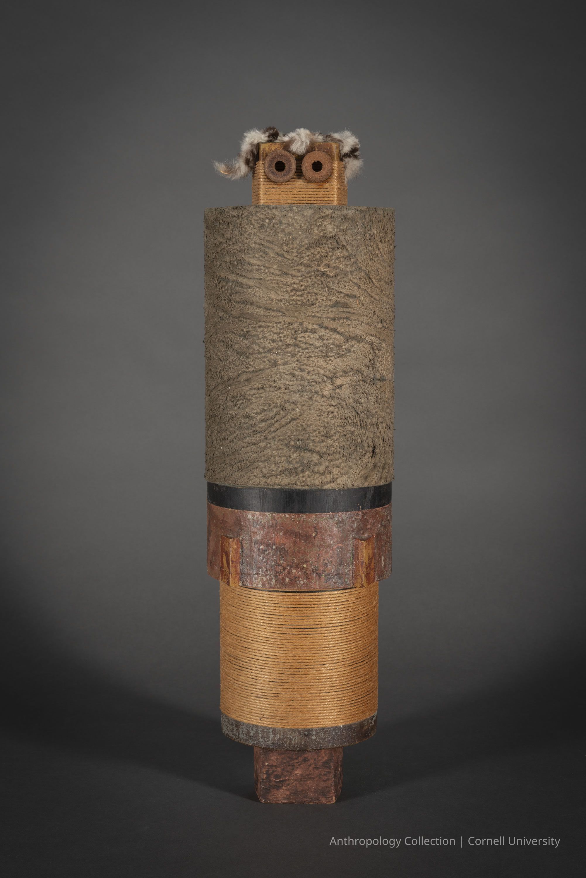 EFFIGY FORM | circa 1966-71 | Photo by Ann Cady, 2024 | ©2026 Norman Daly Art: The photo depicts a vertical cylindrical sculpture composed of various natural materials, including a textured upper section resembling tree bark, a middle portion wrapped in coarse thread, and a lower segment with a rust-like appearance. At the top, a small head-like rectangular structure with two round, eye-like elements, protrudes slightly. it is topped with fluffy, hair-like accents.