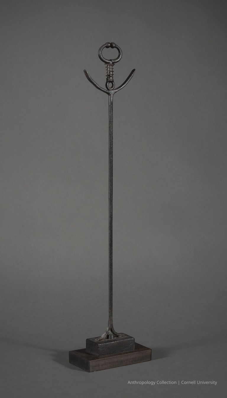 VOTIVE | circa 1961-65 | Photo by Ann Cady, 2024 | ©2026 Norman Daly Art — The photo depicts a minimalist iron artifact featuring a long, slender rod with an elegantly curved, anchor-like top, set upon a rectangular wooden base. The thin metal rod extends upward, culminating in a small, circular loop with two ornate beads perched on either side, close to its apex.