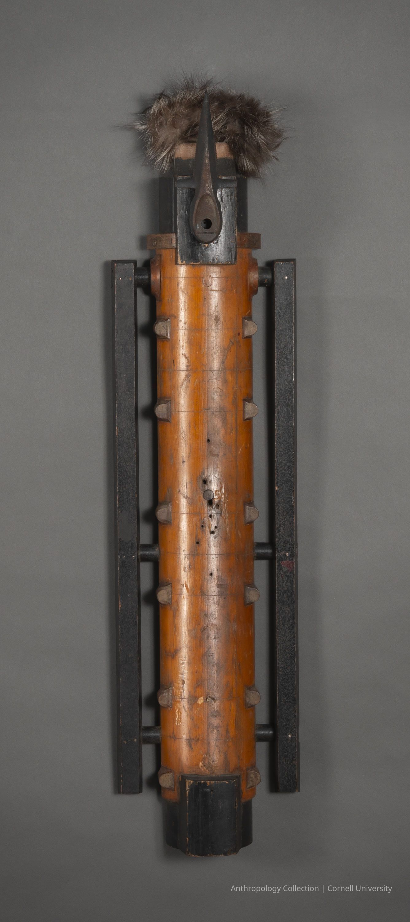 MACE | 1966-71 | Photo by Ann Cady, 2024 | ©2026 Norman Daly Art: A wooden sculpture with an elongated, cylindrical shape, featuring geometric black accents and protruding elements along its sides; the top adorned with a headdress made of dark, textured material resembling fur, and a long, pointed protrusion extends from the upper section, giving the piece an abstract, mask-like appearance, a striking and somewhat mysterious design.