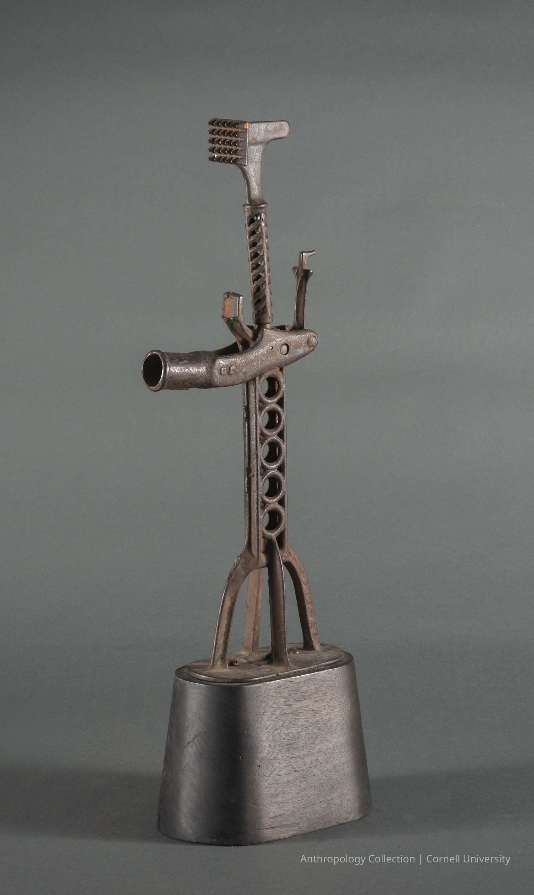 HOME VOTIVE | circa 1961-65 | Photo by Ann Cady, 2024 | ©2026 Norman Daly Art: An artifact composed of various metal components, including a curved, hollow pipe extending from its center, a base with a cylindrical structure, and other intricate metal pieces assembled in a way that mimics a humanoid form; its gray surface is textured and rusted.