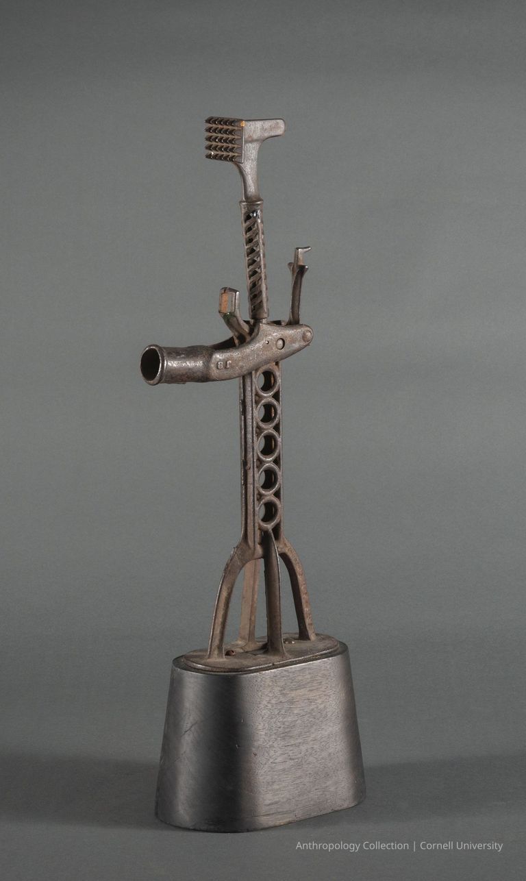 HOME VOTIVE | circa 1961-65 | Photo by Ann Cady, 2024 | ©2026 Norman Daly Art — An artifact composed of various metal components, including a curved, hollow pipe extending from its center, a base with a cylindrical structure, and other intricate metal pieces assembled in a way that mimics a humanoid form; its gray surface is textured and rusted.