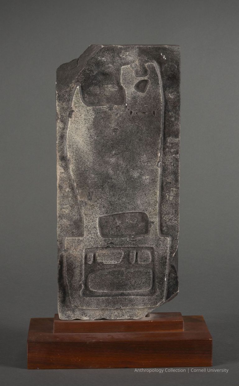 DOUBLE-FACED STELE (side b) | 1960-1965 | Photo by Ann Cady, 2024 | ©2026 Norman Daly Art — The other side of the stele shows an ancient marble artifact featuring engraved symbols and designs, prominently displaying geometric shapes and what appears to be script. It is mounted on a polished wooden stand against a gray background, giving prominence to its historical and archaeological significance. The artifact’s texture and wear suggest significant age, likely making it an important piece for study in cultural or historical contexts.