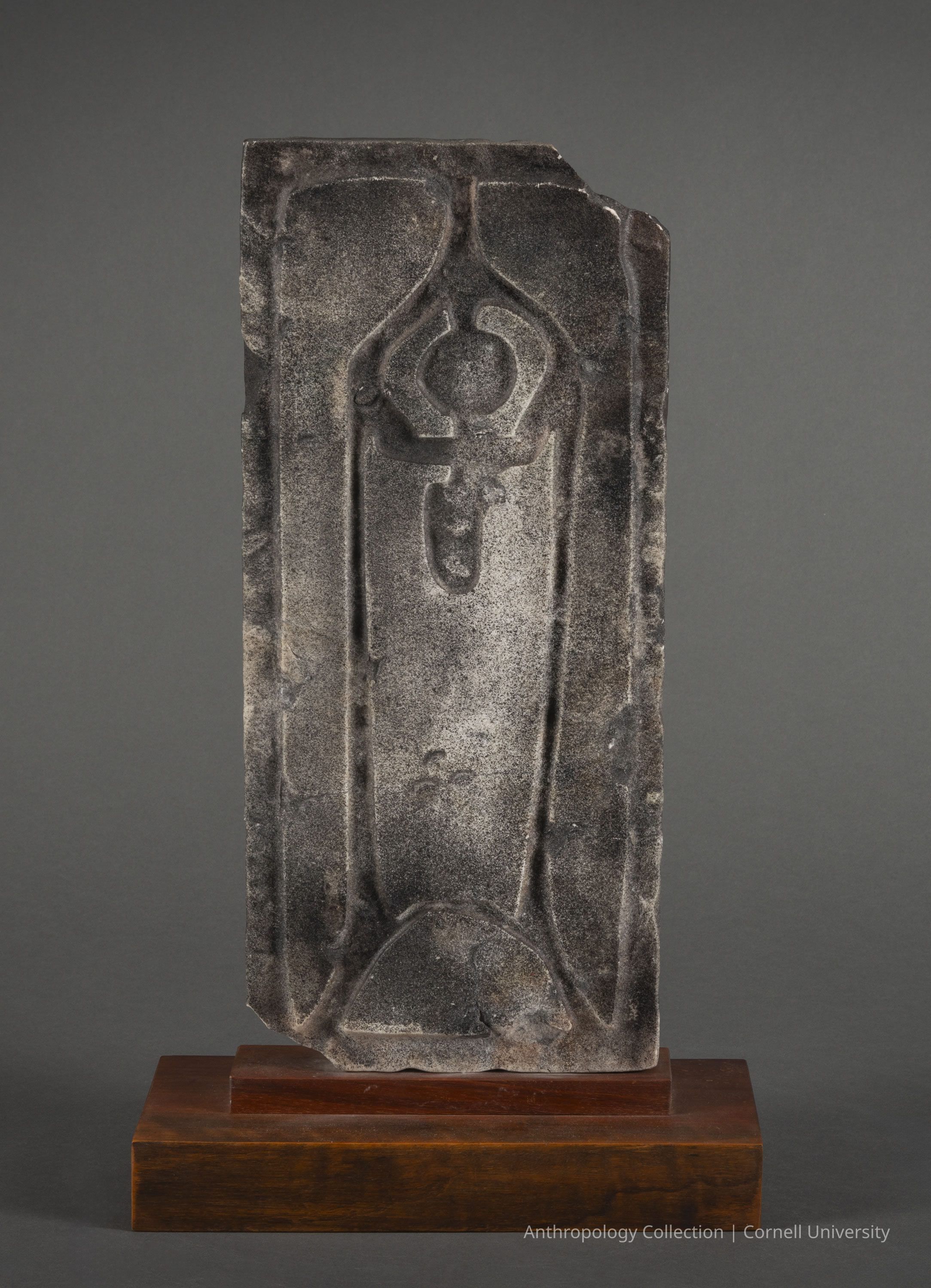 DOUBLE-FACED STELE (side a) | 1960-1965 | Photo by Ann Cady, 2024 | ©2026 Norman Daly Art: A weathered, rectangular slab of marble featuring a deeply carved, symmetrical design supported by a polished wooden base. The central motif is a vertical, elegantly shaped orant figure surrounded by distinct, curvilinear patterns that suggest a ceremonial purpose. The stone's rough texture contrasts with the meticulously crafted lines and shapes, creating an intriguing visual archeological piece. The gray backdrop highlights the sculpture’s antiquity and aesthetic form.