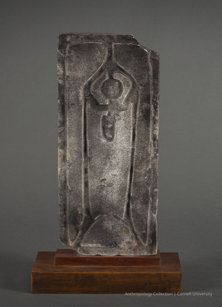 DOUBLE-FACED STELE (side a) | 1960-1965 | Photo by Ann Cady, 2024 | ©2026 Norman Daly Art — A weathered, rectangular slab of marble featuring a deeply carved, symmetrical design supported by a polished wooden base. The central motif is a vertical, elegantly shaped orant figure surrounded by distinct, curvilinear patterns that suggest a ceremonial purpose. The stone's rough texture contrasts with the meticulously crafted lines and shapes, creating an intriguing visual archeological piece. The gray backdrop highlights the sculpture’s antiquity and aesthetic form.