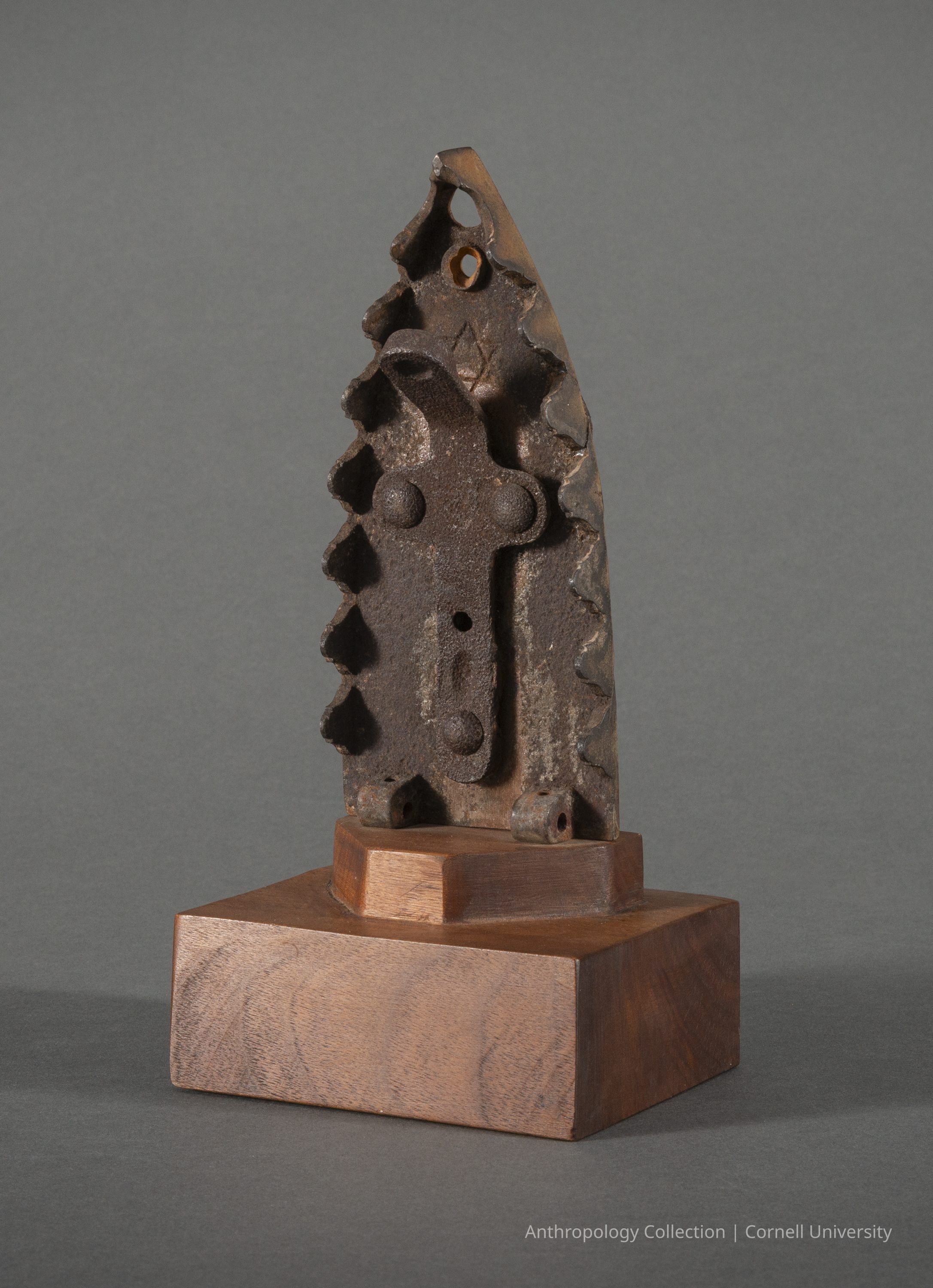 VOTIVE | 1961-65 | Photo by Ann Cady, 2024 | ©2026 Norman Daly Art: The photo features an archaic iron artifact, resembling a flat iron, placed vertically on a wooden base. The central part of the metal object displays a prominent cross with circular detailing and additional geometric cut-outs. Two pierced holes at the top apex and the middle part of the cross suggest a decorative or functional purpose. The object exhibits a weathered surface with rust and wear, indicating age and historical use. The two-tiered base added later shows a polished wood construction.