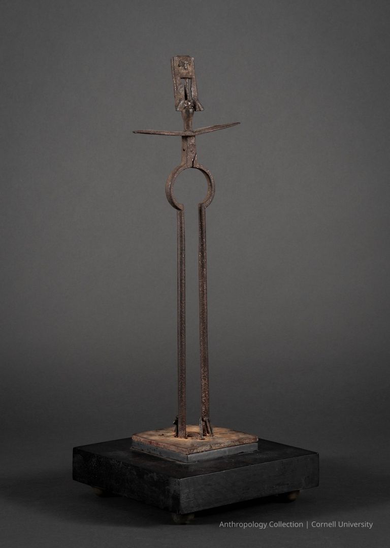 VOTIVE FIGURE | 1965 | Photo by Ann Cady, 2024 | ©2026 Norman Daly Art — This image displays an evocative metal sculpture composed of various iron elements, including an elongated vertical shaft topped by a stylized humanoid figure with extended arms resembling a crucifix. The structure is mounted securely on a square wooden base, highlighting the rustic texture and aged quality of the materials utilized. The overall impression is one of stark, simplified form, merging elements of the abstract with symbolic religious iconography. The dark backdrop accentuates the sculpture's silhouette and the interplay of light on the textured metal.