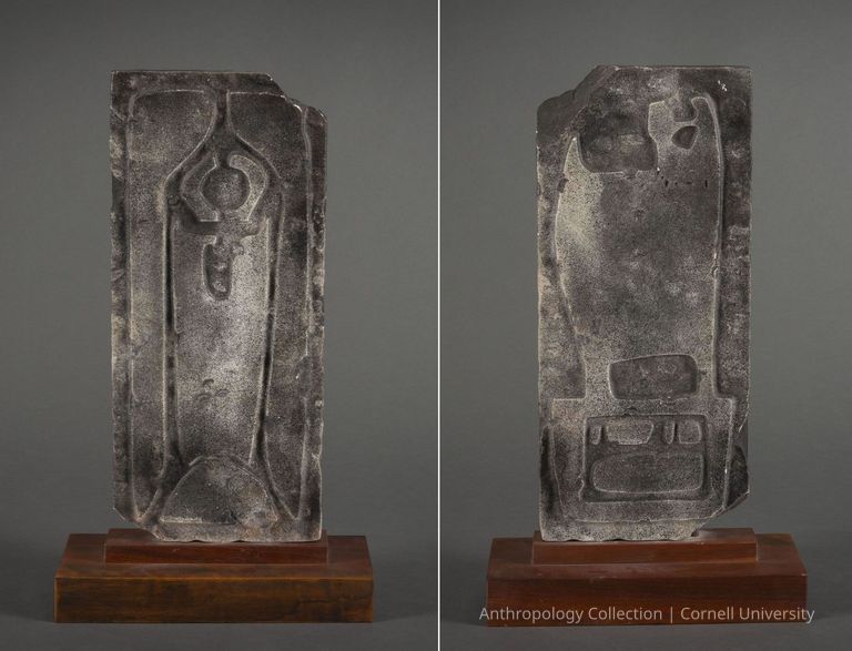 DOUBLE-FACED STELE | 1960-1965 | Photos by Ann Cady, 2024 | ©2026 Norman Daly Art — A weathered, rectangular slab of marble featuring a deeply carved, symmetrical design supported by a polished wooden base. The central motif of the left face is a vertical, elegantly shaped orant figure surrounded by distinct, curvilinear patterns that suggest a ceremonial purpose. The other side of the stele features engraved symbols and designs, prominently displaying geometric shapes and what appears to be script. The stone's rough texture contrasts with the meticulously crafted lines and shapes, creating an intriguing visual archeological piece.