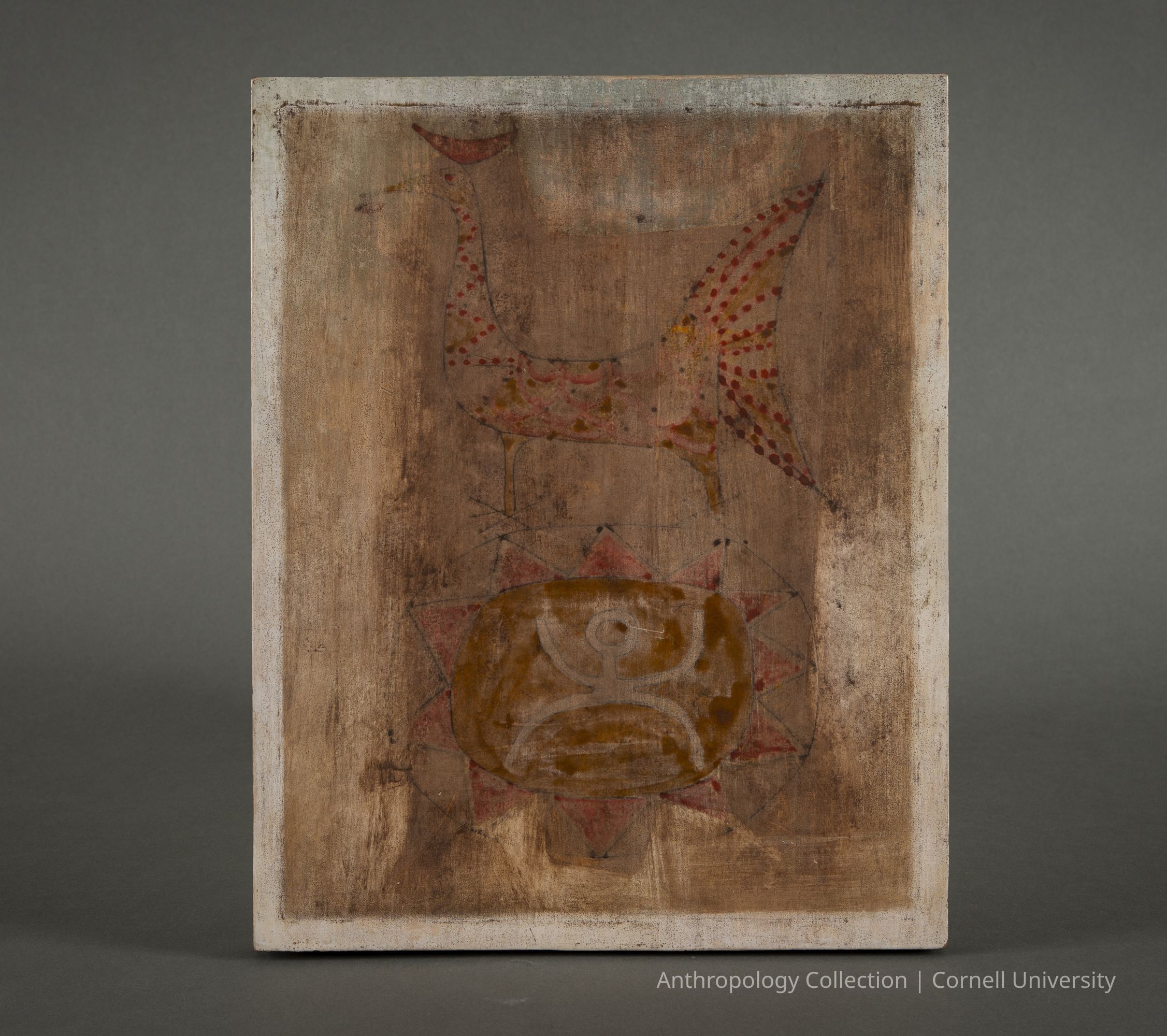 Facsimile of Wall Painting with Stilt-Walker and Speckled Bird | circa 1967-71 | Photo by Ann Cady, 2026 | ©2026 Norman Daly Art: A rustic, textured wooden panel featuring a combination of muted earth tones and red accents as background. At the top, an elongated form resembling a stylized, mythical bird-like creature is adorned with red circular patterns that accentuate its shape. Beneath it, a rounded, intricate symbol with swirling elements is enclosed within a star-like motif, the points of which extend outward, adding to the visual interest and complexity. A figure with upraised arms is situated at the center.