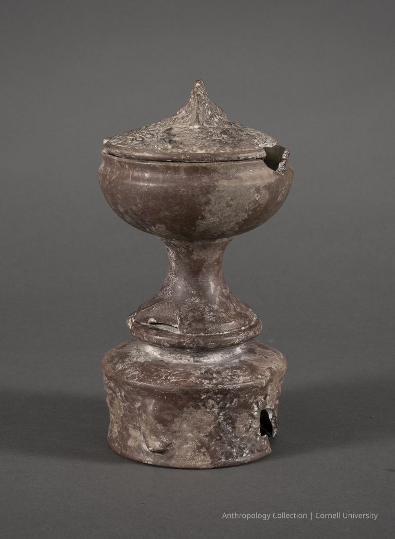 URN | circa 1966-1971 | Photo by Ann Cady, 2024 | ©2026 Norman Daly Art — An antique ceramic urn with a delicately detailed lid, featuring floral embellishments and a pointed top. The artifact has a distinguished pedestal base with a rounded, bulbous stem leading to a wide foot that appears lightly damaged or eroded, indicating considerable age and wear. The surface of the urn has a mottled patina that adds to its historical aura, suggesting it might be a relic used in ceremonial contexts.