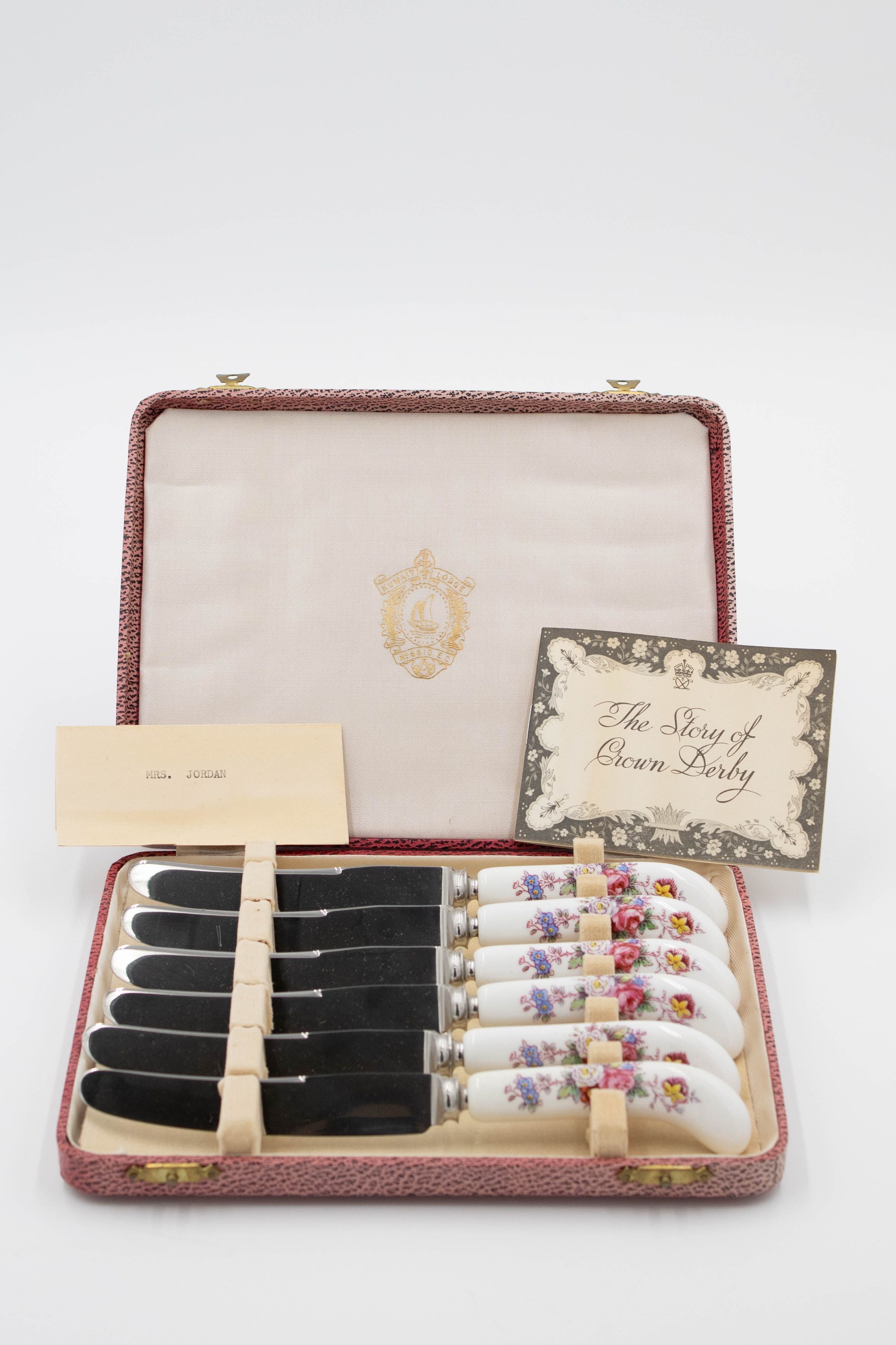 Set of Royal Crown Derby knives given to Mrs. Jessie Jordan.: Image of the set of six knives decorated with a Royal Crown Derby posies pattern. They are in a red leather case with a cream felt interior. There are two cards, the left one reads "MRS. JORDAN" and the right one reads "The Story of Crown Derby." On the inside of the lid, there is a gold printed image of the seal of the Kuwait Masonic Lodge. The seal has a Kuwaiti dhow in the middle and the traditional Masonic compass at the center bottom.