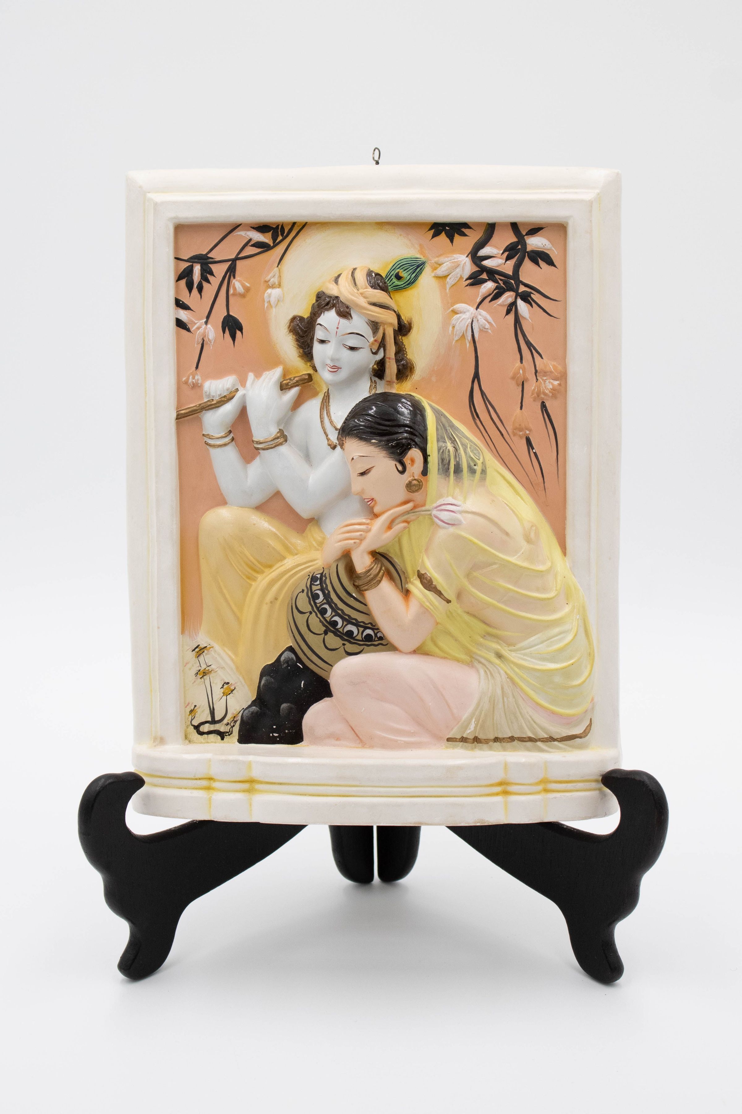 Molded ceramic picture painted with Radha and Lord Krishna, likely from India.: Image of the molded ceramic picture painted with Radha and Lord Krishna on a black picture stand. Radha is depicted in light pink clothing with a yellow shawl draped over her head. Krishna is seen behind Radha and is playing a flute with a peacock feather in his hair.