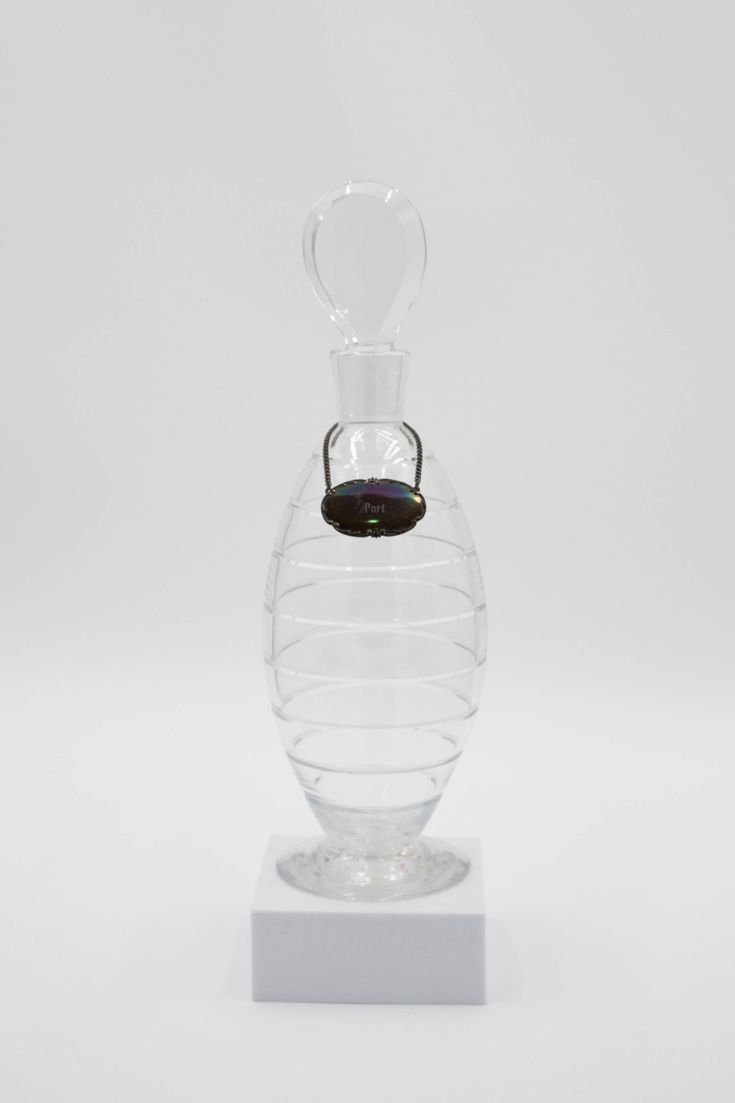 Crystal port decanter: A glass decanter with a small silver plaque that says "Port" and sits on the decanters neck like a necklace. The decanter has a flat base and is considered a standard shaped decanter. There are opaque horizontal striped going up its oval shaped body. The decanter has a short neck and a round, bulb shaped stopper.
