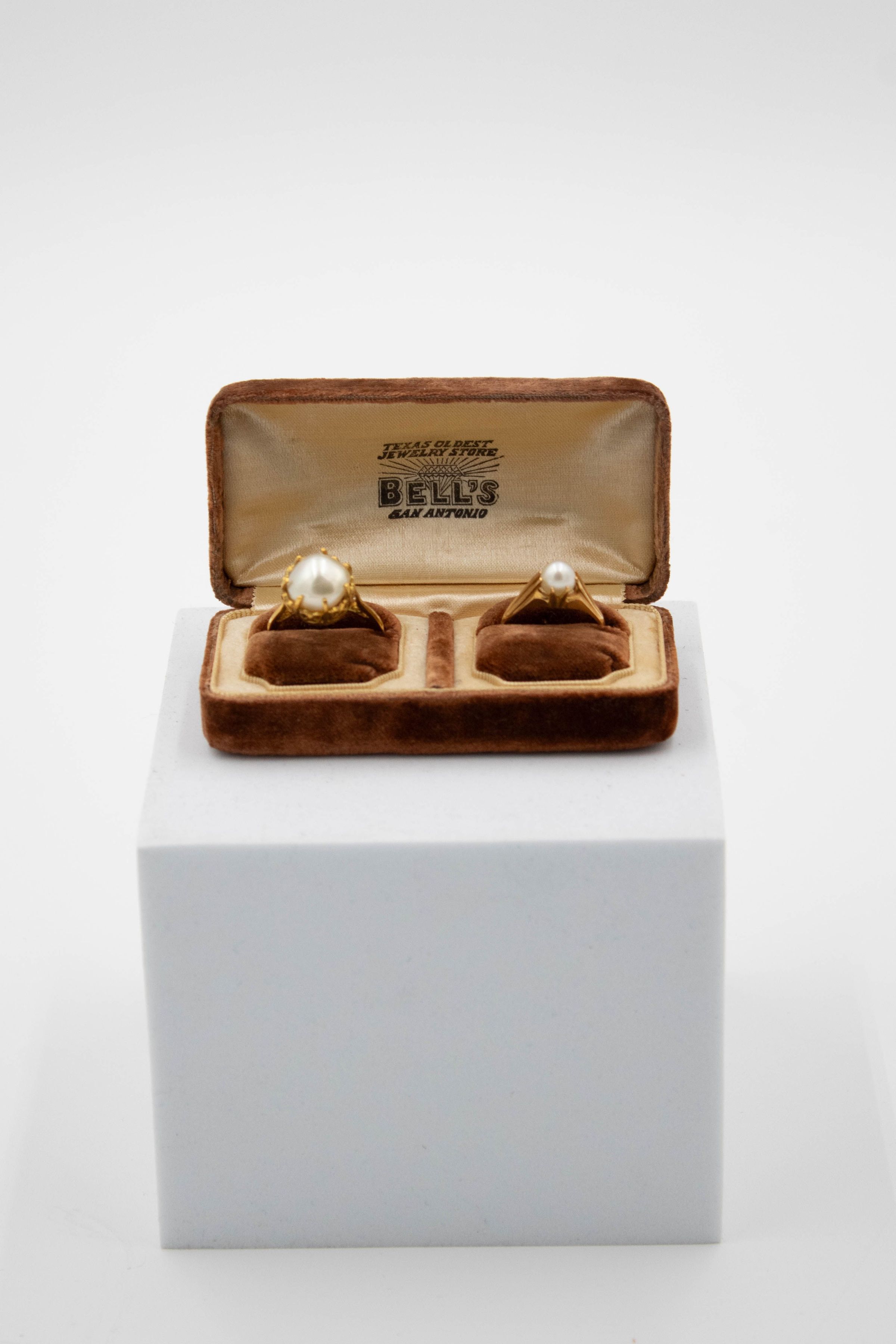 Pair of gold and pearl rings in a "Bell's San Antonio" double ring box.: Image of double brown felt ring box with a pair of gold and pearl rings. Each of the rings sit on a brown felt pillow and the inside of the box is yellow satin. The inside lid reads "TEXAS OLDEST JEWELRY STORE BELL'S SAN ANTONIO" in black letters. The ring on the left has a larger pearl, prong setting, and a narrow band, while the one on the right has a smaller pearl with a wide, tapered band.