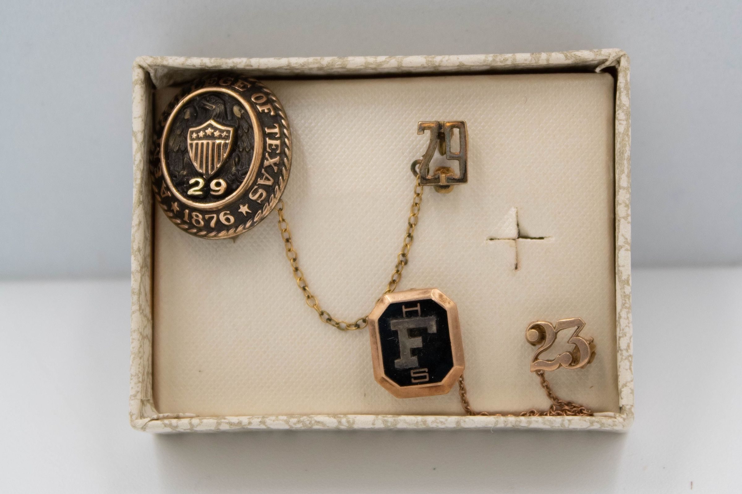 Up close image of L.T.'s class pins from Texas A&M University and Floresville High School in a marble-colored cardboard box. The Texas A&M pin a gold oval with an eagle and shield in the center. The number "29" is under the shield, denoting L.T.'s class year. The words "A&M COLLEGE OF TEXAS 1876" is surrounding the outside of the oval. There is a gold chain that attaches to the "29" on a separate pin. The Floresville High School pin is in a similar fashion but it is a rectangle. It is bronze with a large "F" in the center. There is a gold chain that attaches to the "23" on a separate pin.