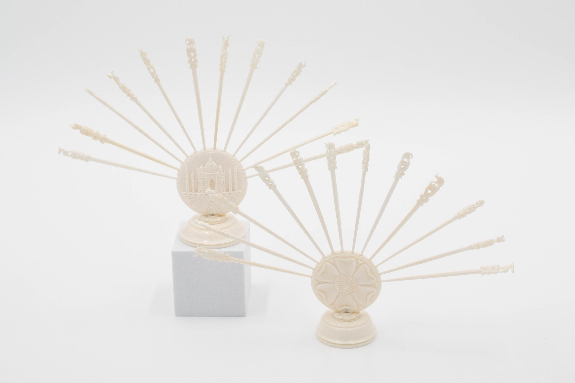 Set of two ivory skewers from India.: Image of the two sets of ivory skewers. The one on the front right has a flower on the base and 12 skewers across the top. The one on the back left has the Taj Mahal on the base and 12 skewers across the top. Each of the skewers either has an elephant or elk on the top.