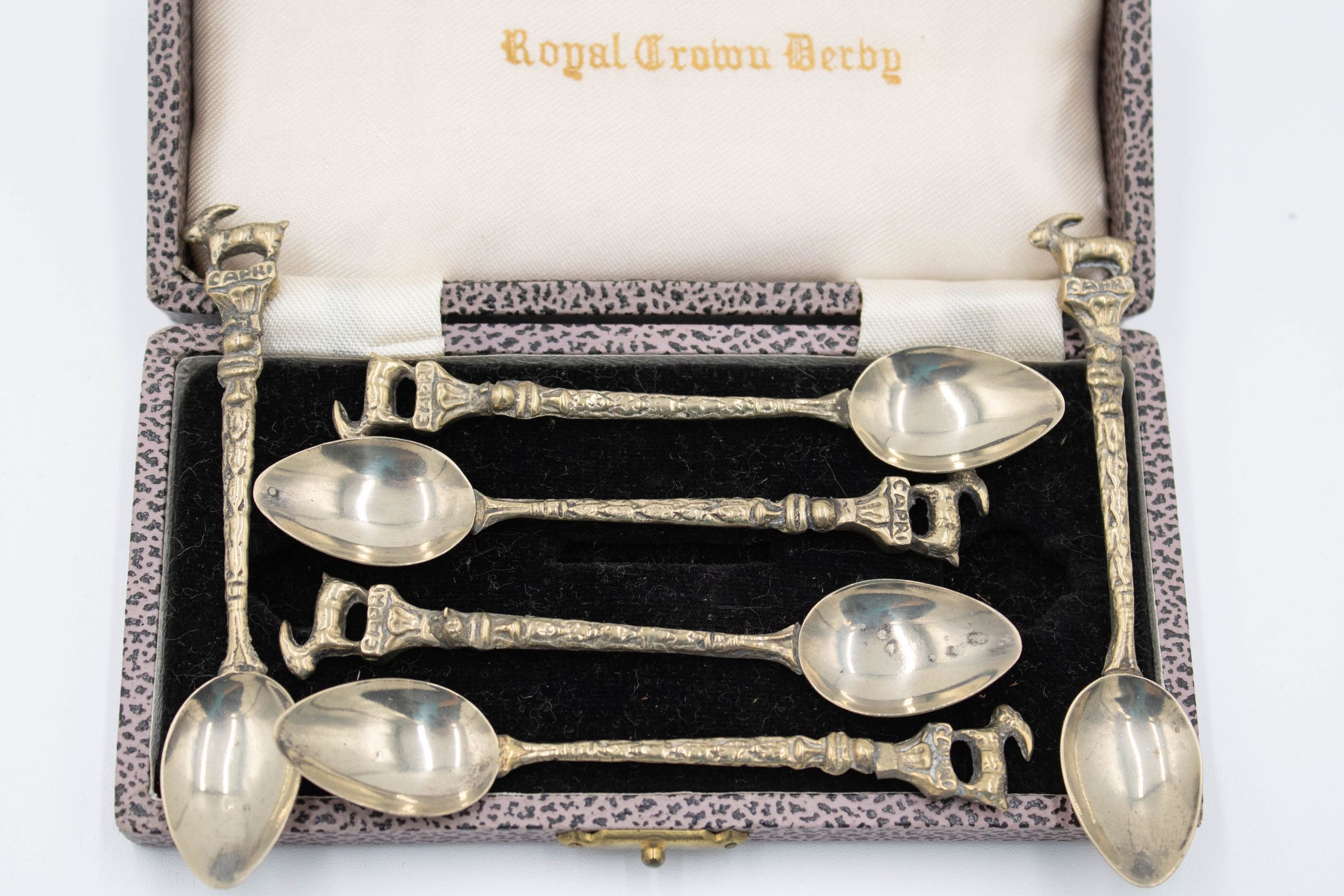 Set of Spoons with Case: Image of all six spoons displayed against the interior of the case. The inside of the case is lined with pink tiger print on its outline, while the bottom part has black felt. There are six divots inside the black felt for the spoons to be held in. The top of the case's interior is lined with a white material. In the middle of the white material are the words "Royal Crown Derby" in a yellow gothic script.