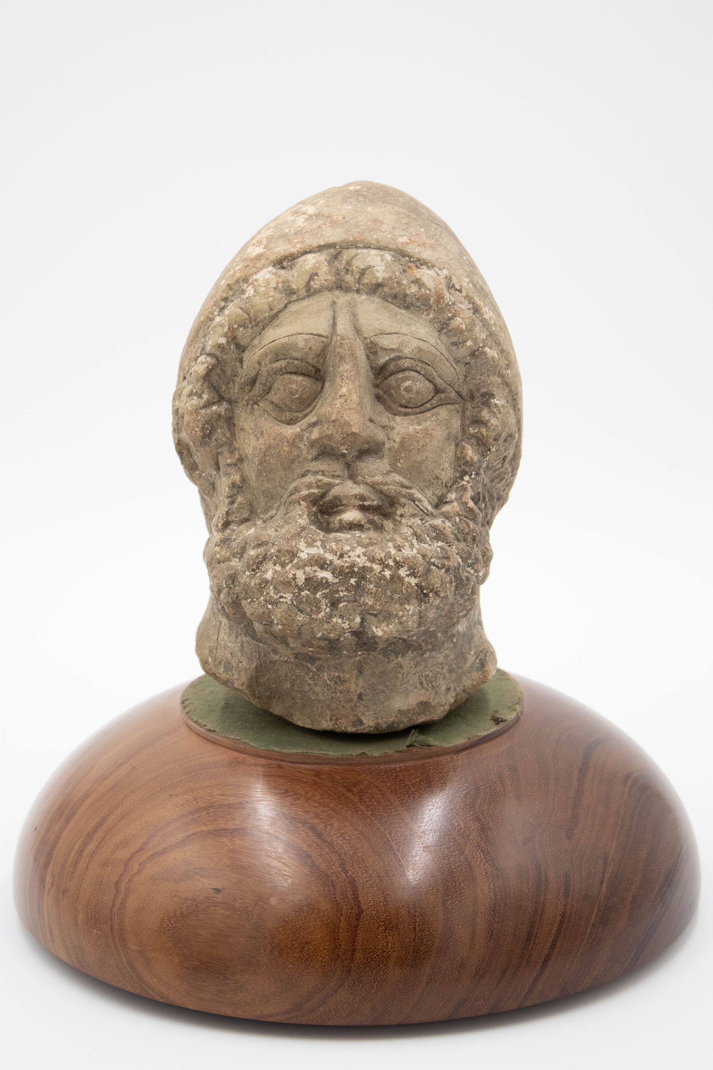 Stone Roman head: A gray Roman man's head carved out of stone. He has curly hair and a beard, with a pointed helmet covering most of his hair. His eyebrows are thin, his eyes are wide, and his nose has visible wear. He sits on a wooden base that is a half-sphere with a flat side for the head to sit on.