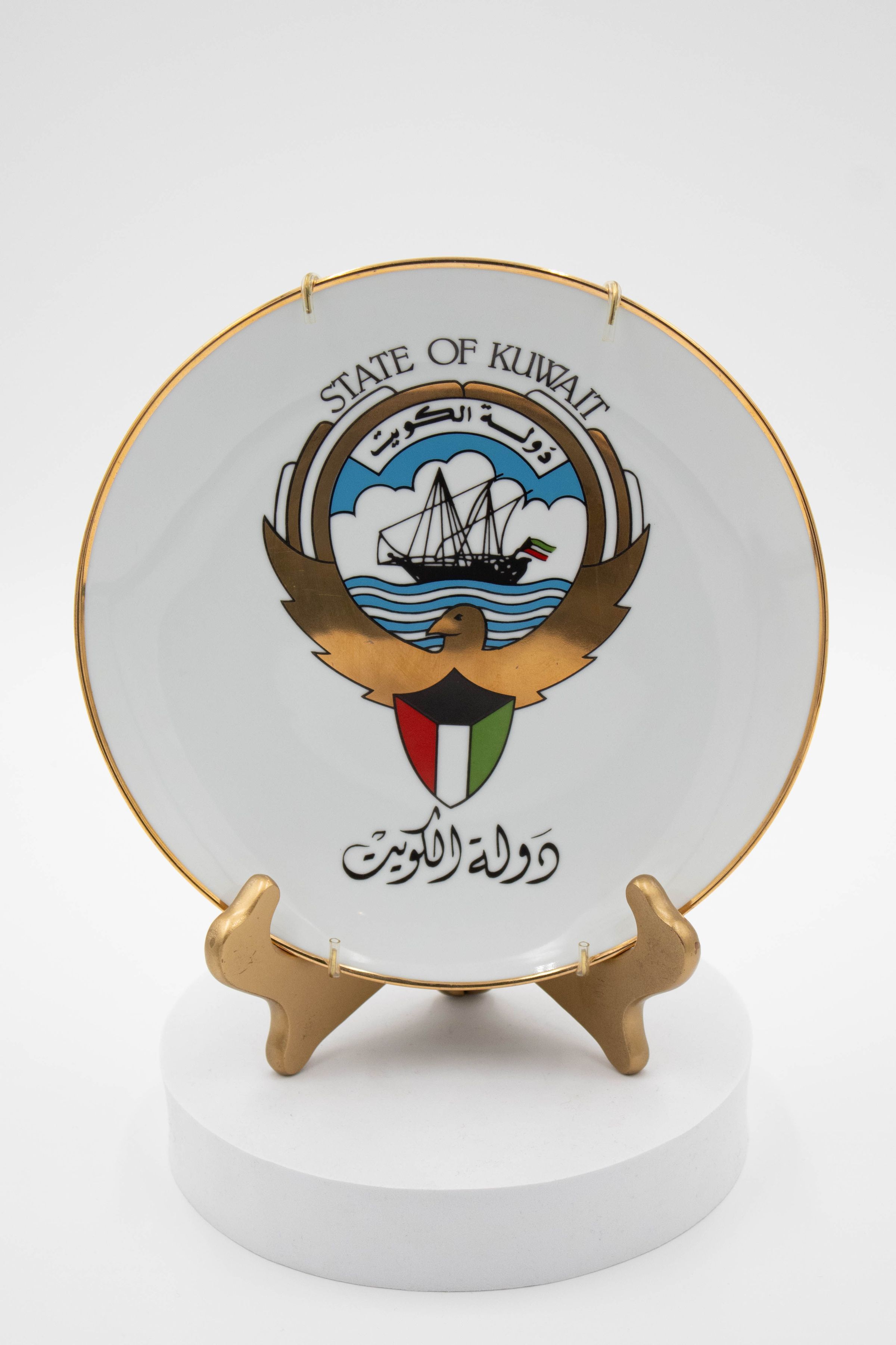 Kuwait Plate on Stand: Image showing the porcelain plate on a gold stand. The plate features a gold rim and showcases the emblem of Kuwait. A Kuwaiti Dhow, flag and falcon make up the emblem's composition. Above the emblem is "State of Kuwait." Below the emblem is "دَوْلَةُ ٱلْكُوَيْت" which is also "State of Kuwait" but in Arabic.