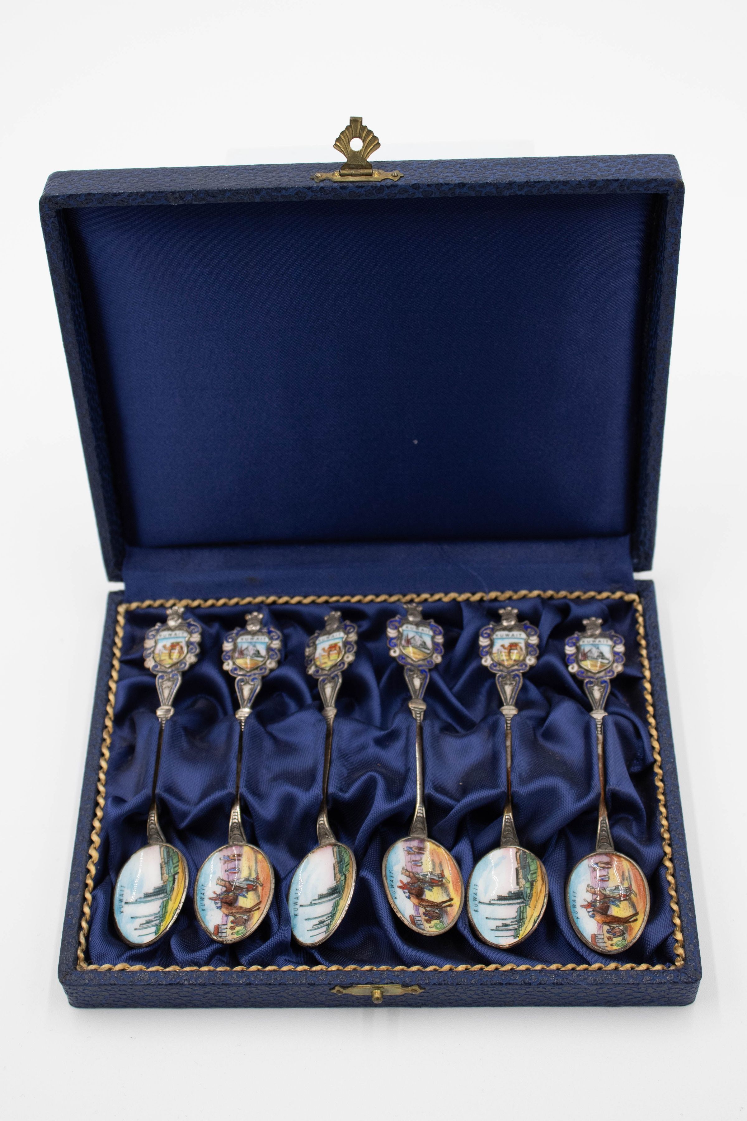 Set of six silver spoons decorated with scenes of Kuwait in a blue case.: Image of the set of six silver spoons in a blue felt case. There are two different designs enameled on both the handle and bowl. One spoon has a camel and Bedouin on the handle with a scene of an oil rig system on the bowl. The handle is also decorated with small silver details and there is a knight helmet on the end. The other design has an oil ship on the handle with camel and Bedouin man in the desert on the bowl. The six spoons are in a blue case.