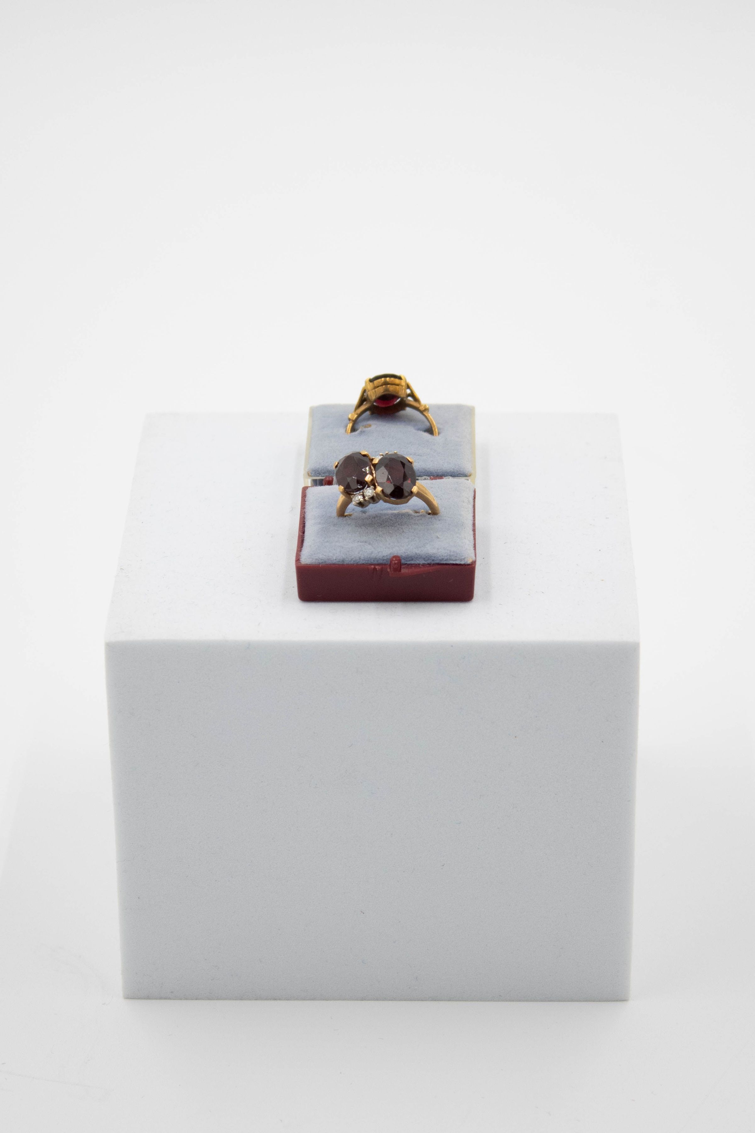 Pair of gold and garnet rings that belonged to Mrs. Jessie Jordan.: Image of the pair of gold and garnet rings with one facing the front. The ring in the front has two oval stones with four small diamonds in a pronged setting on a thin gold band. The rings are in a double sided box with blue cushions and a maroon plastic box.