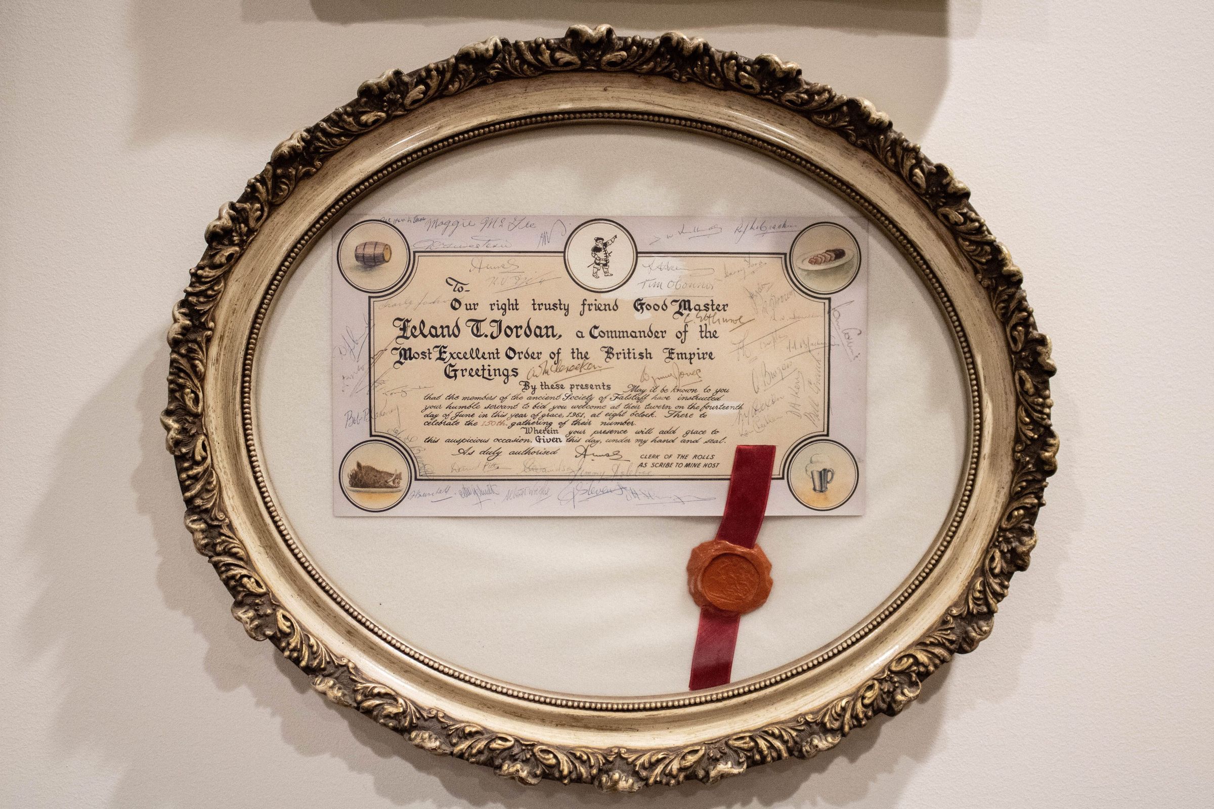Certificate in Frame: Image of the oval frame which encases the certificate. The frame is ornate and has decorative designs. The material is a white wood and appears to be dirty. The certificate is off-center, with the bottom of the frame's open white space occupied with a red ribbon and orange wax seal.