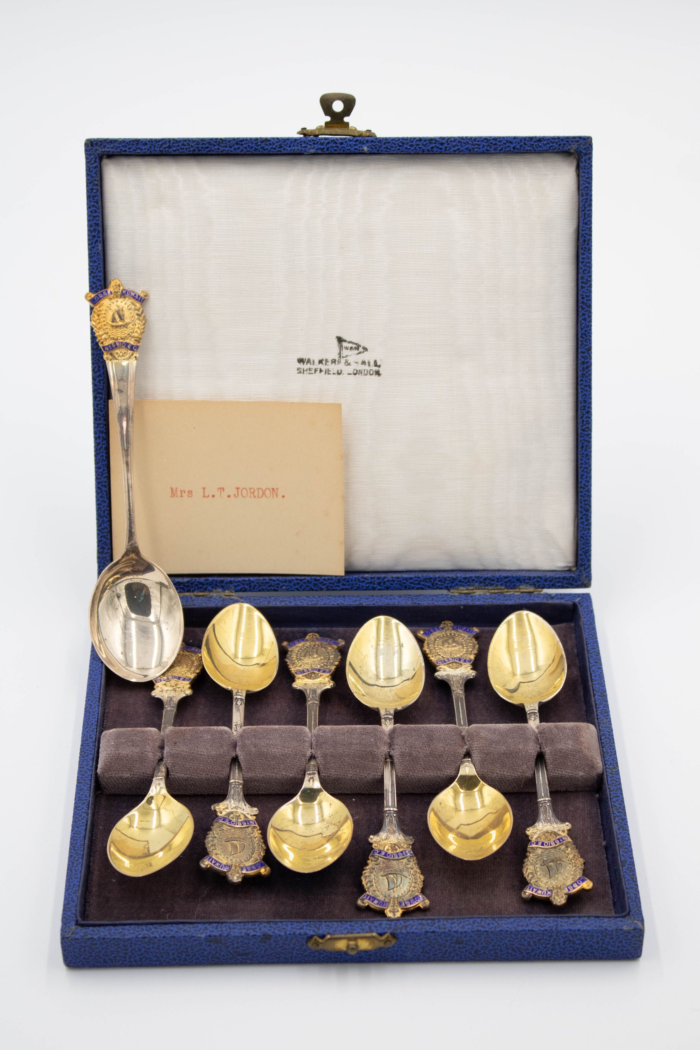 Set of Kuwaiti Coffee Spoons in Blue Case: Image of the spoons in their case. The case is open and is made of a blue leather with a brass lock. It has a black leopard pattern. There are six coffee spoons laying in the case while one soup spoon is featured upright. Each spoon has a Masonic Crest ornament on each of their handles. The soup spoon's bowl is silver and oblong while the coffee spoons' bowls are gold. There is a weathered yellowed note in the case.