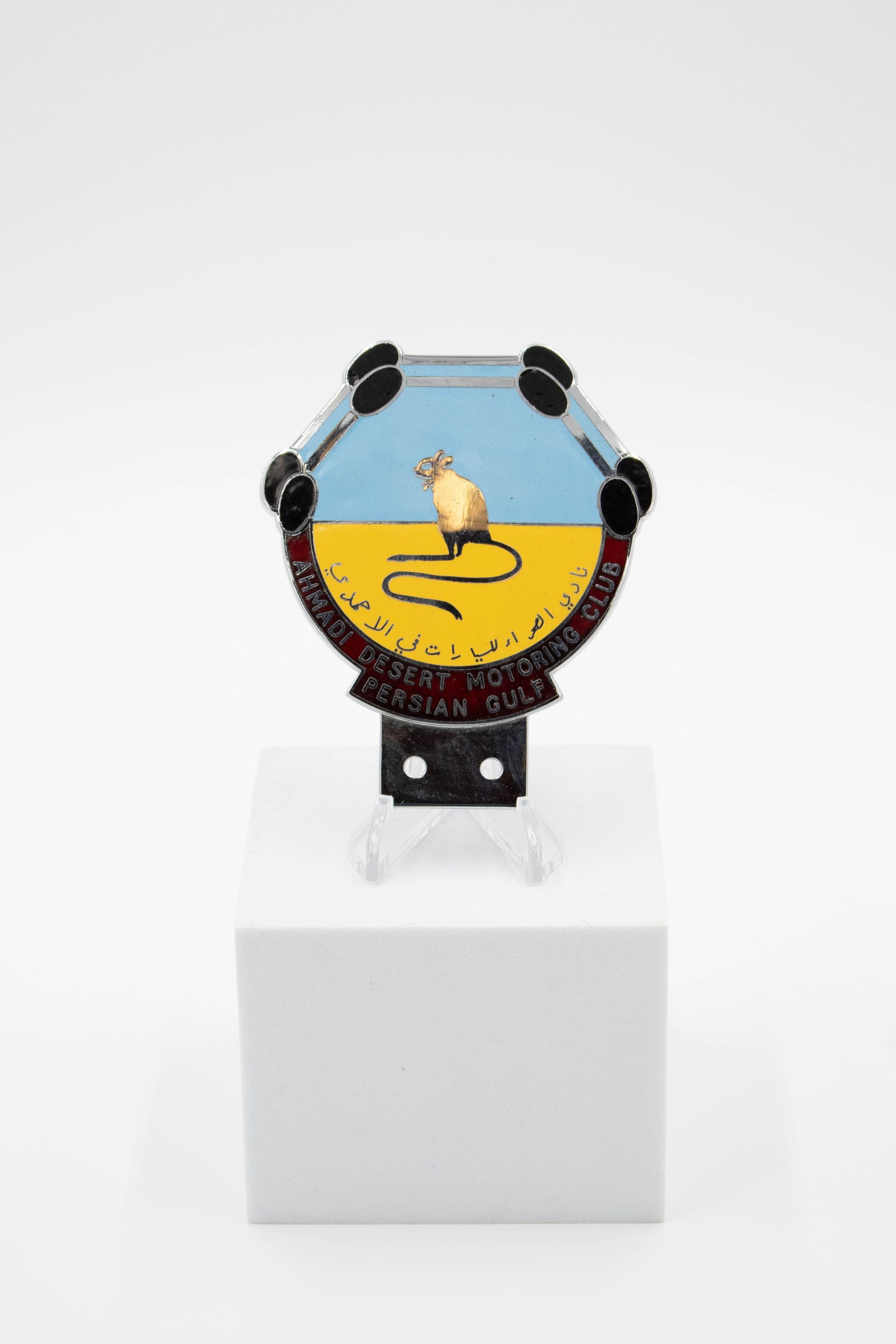 Ahmadi Desert Motoring Club Car Badge: A palm-sized blue and yellow car badge. It is yellow and half-circular on the bottom, with four trapezoidal edges on the blue top. Around the bottom edge, it says "Ahmadi Desert Motoring Club Persian Gulf." A row of Arabic text states the car club's title. In the center of the badge a rodent is featured with long lower legs, a long tail and small ears, likely a jerboa. It weighs 194 grams and is 4.5 inches tall, 4 inches long and is roughly 1/8th of an inch thick.