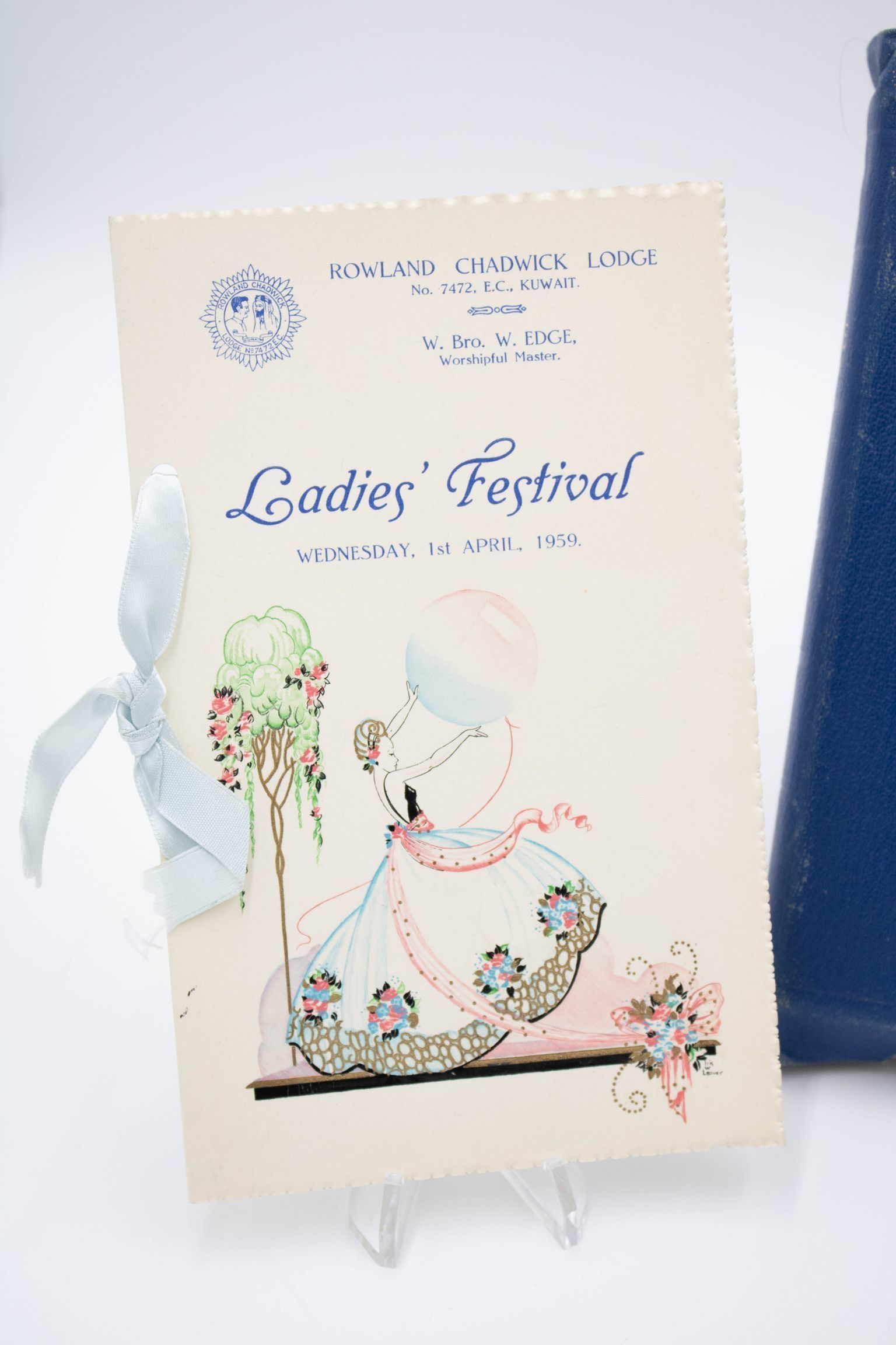 Program from the Ladies' Festival at the Kuwait Masonic Lodge.: Image of the cream-colored program with woman in long dress and balloon on the front. The binding is a baby blue ribbon. The front of the program reads "ROWLAND CHADWICK LODGE No. 7472, E.C., KUWAIT" on the top with "W. Bro. W. EDGE, Worshipful Master" under. Additionally the program says "Ladies' Festival WEDNESDAY, 1ST APRIL, 1959."