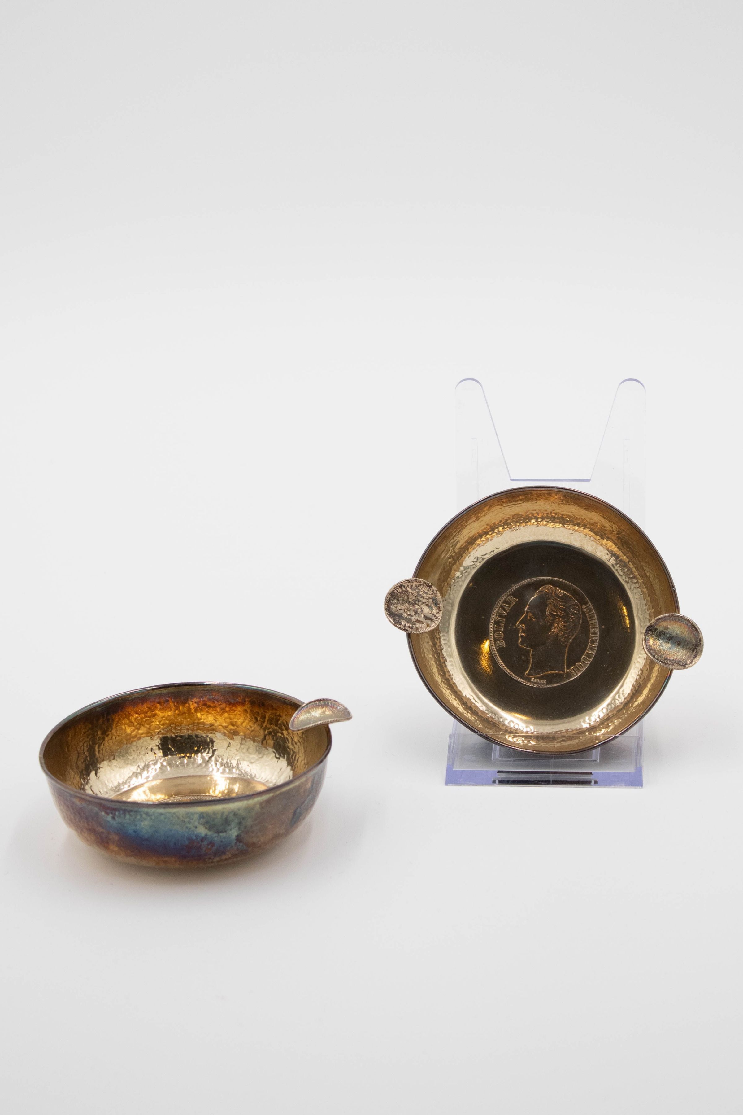 Pair of Small Brass Ashtrays: Image of the two brass ashtrays. The one on the right is on a plastic stand which reveals that the bottom of the ashtray has a Bolivar stamped into its center. The coin takes up much of the bottom and its obverse depicts a bust of Simon Bolivar looking to his left, with the word "Bolivar" on the left and "Libertador" on the right. The two cigarette holders are made out of bent bolivars. The other ashtray is a similar size but has only one cigarette holder.