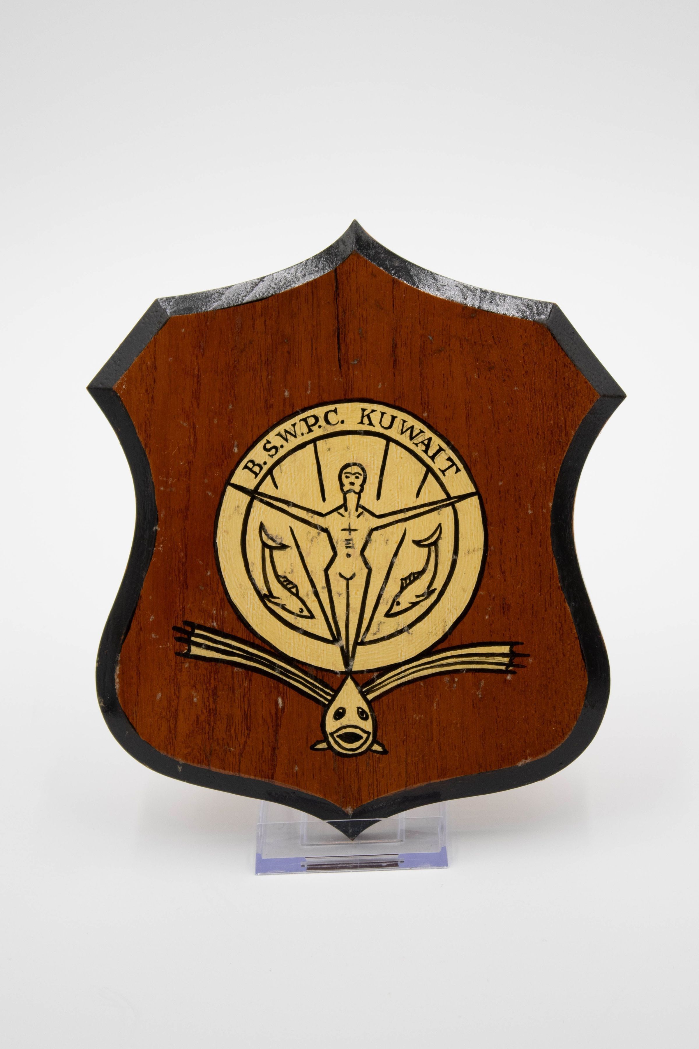 B.S.W.P.C. Kuwait Field Hockey Club Plaque with Man and Fish: A dark, red-tinted plaque in an irregular shape. It has a gold circle with a simply drawn man. His arms are up and out in a proud pose. There are two fish near his legs and a forward-facing fish with big fins at the bottom. The top of the plaque says "B.S.W.P.C KUWAIT." It is 8 inches from top to bottom.