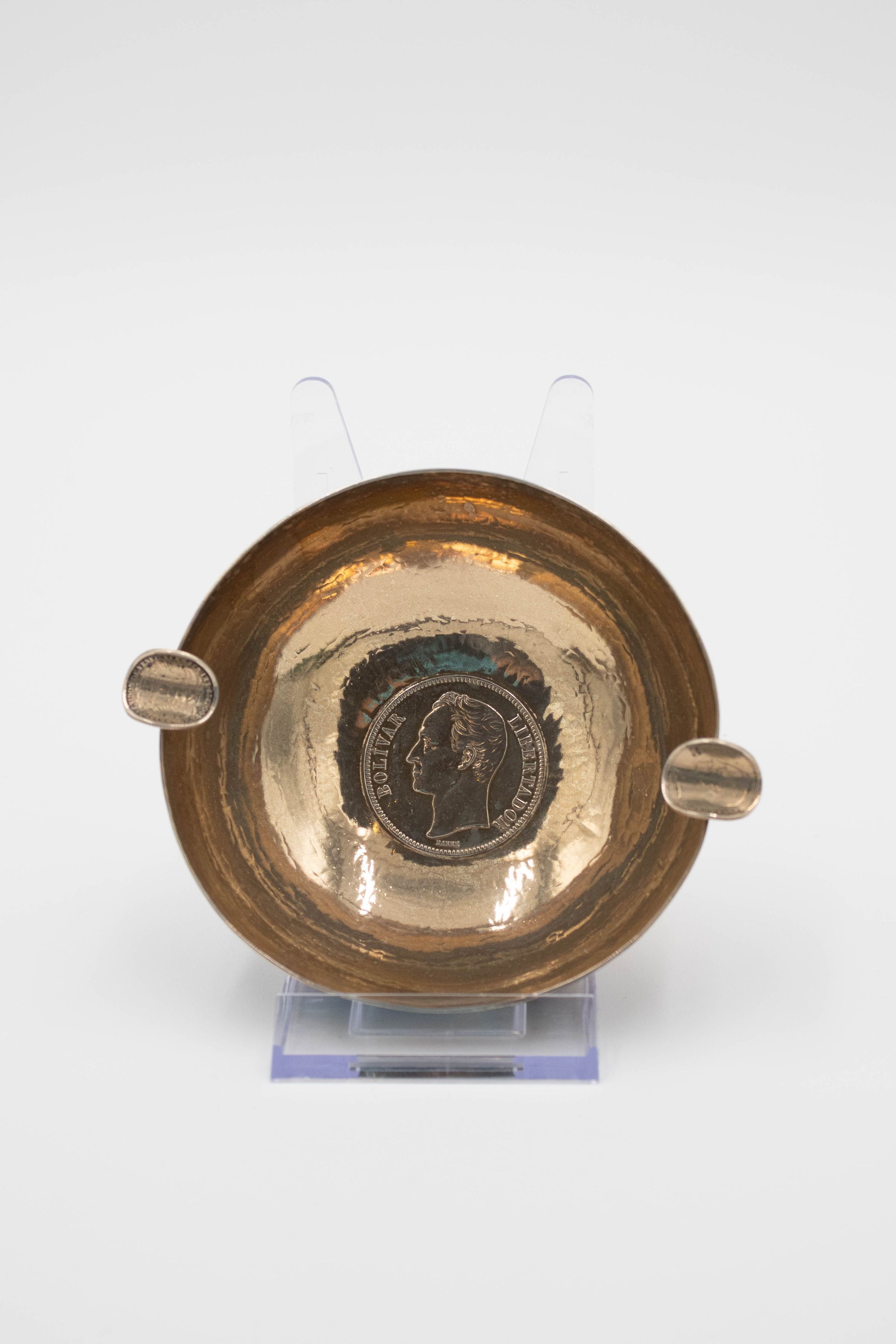 Brass Ashtray: Image of the brass ashtray on a plastic stand to have the interior be visible. The bottom of the ash tray has a Venezuelan Bolivar stamped into its center. The coin depicts the bust of Simón Bolívar facing left with "Bolívar" on the left of the coin and "Libertador" on the right. "Barre" is below Bolívar's bust. There are two cigarette holders on each end of the ashtray, each made with bent Bolivars.