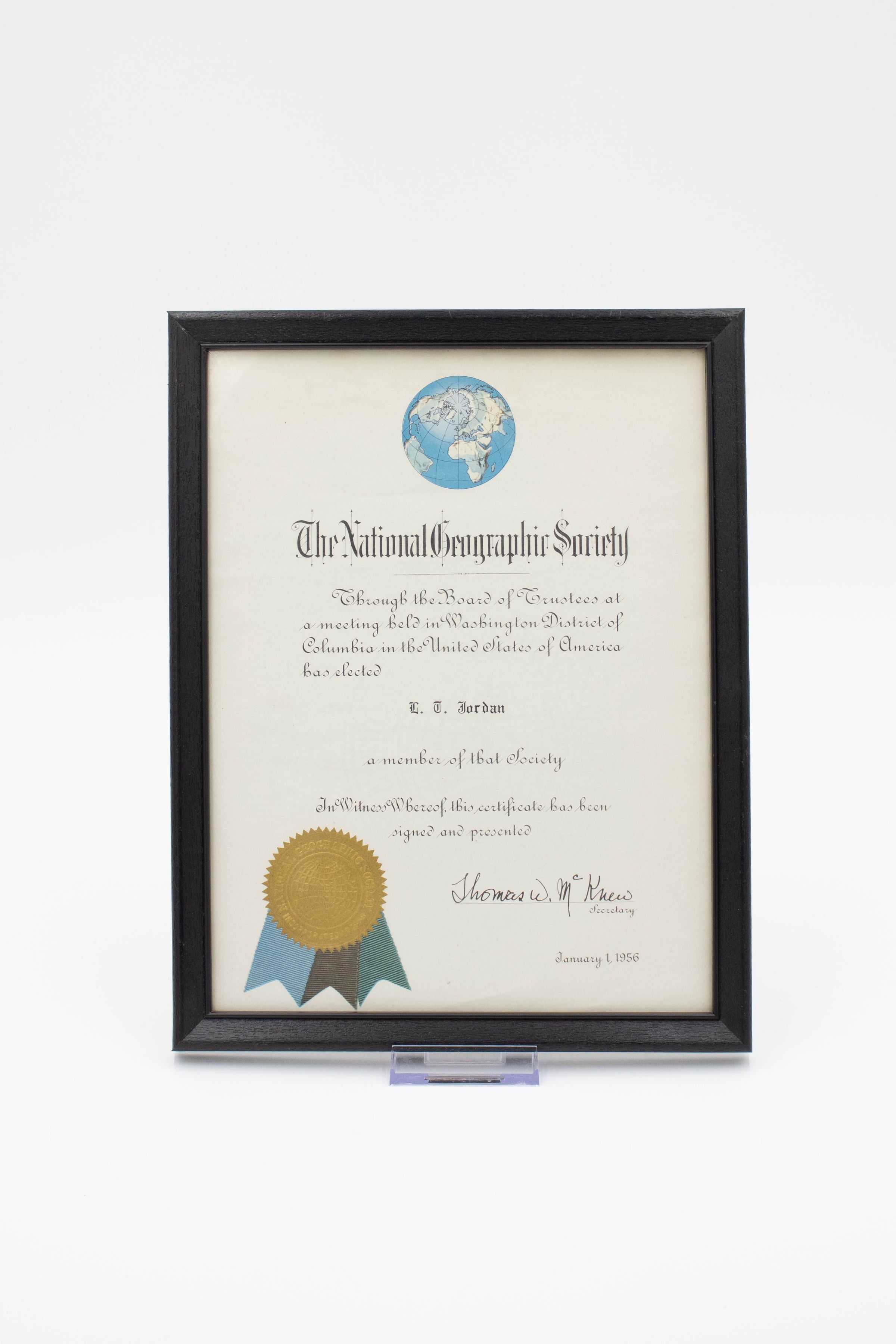 Certificate in Frame: Image of the certificate in its black wooden frame. The top of the certificate features a depiction of a blue globe from a top-down perspective fixated on the north pole. The bottom left of the certificate has a gold seal from the National Geographic Society, with three short ribbons in three shades of blue.