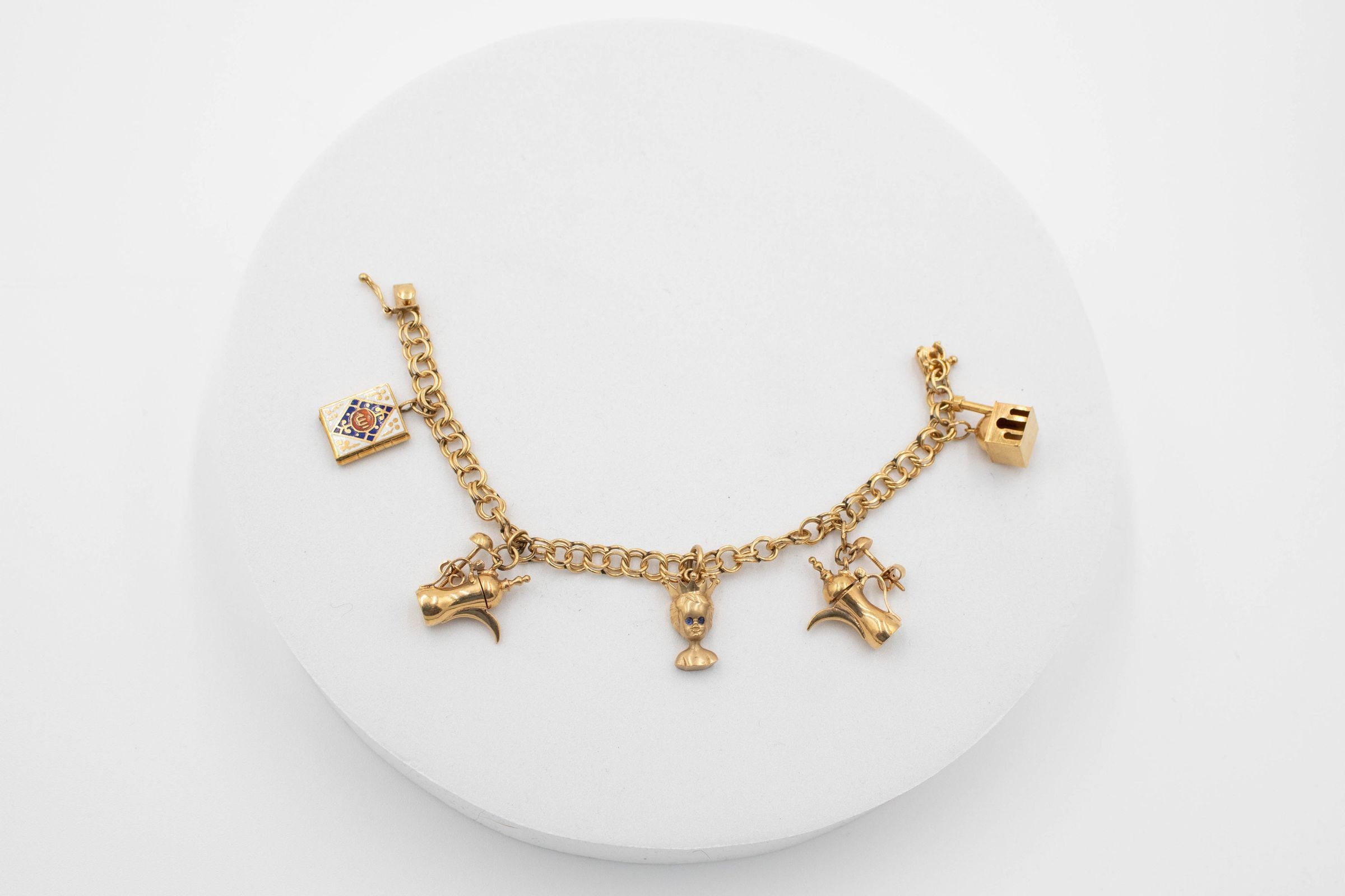 Gold five-charm bracelet that belonged to Mrs. Jessie Jordan.: Image of the gold five-charm bracelet on gold chain. There is a book charm with Arabic script, two small Arabic coffee pots, a small mosque building, and a bust of a boy with a crown and blue gem eyes. The two coffee pot charms are earrings and still have the mechanisms attached to hang them. The bracelet bracelet is Texas barbed wire-style and clasp is a four-leaf clove.