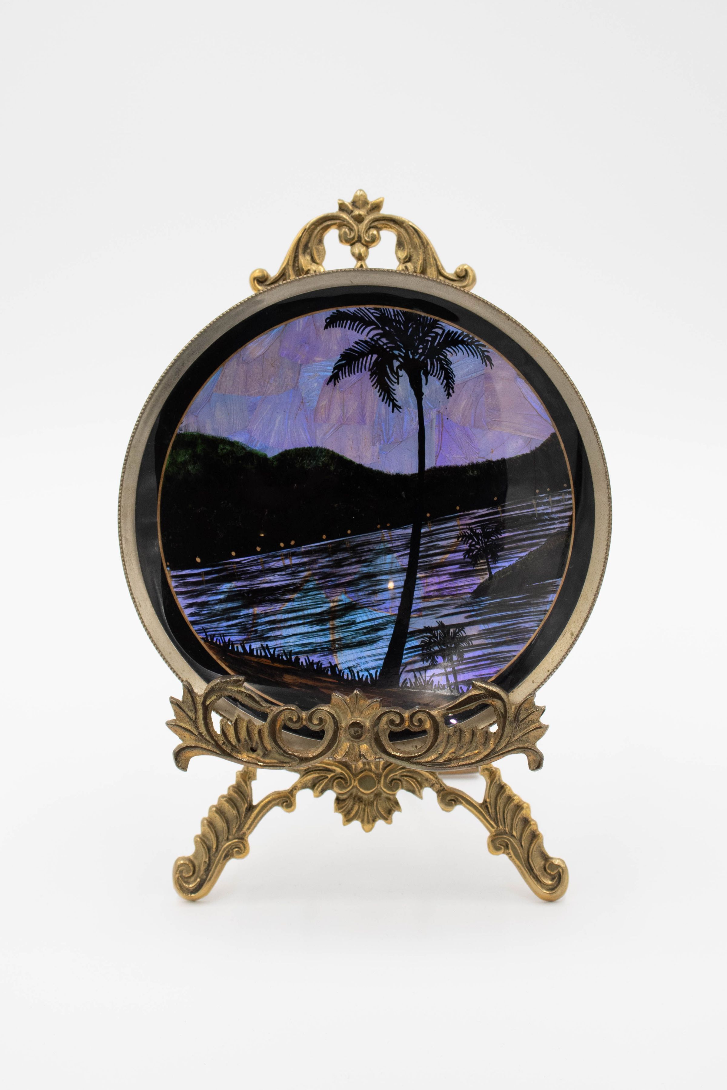 Oasis Scene Plate on Gold Stand: Image of the plate on its gold stand. Painted on the plate is an oasis, with a palm tree in the foreground and a body of water and mountains in the background. The scene is colored using blue, purple, and black from butterfly wings. The stand is a lusterless gold and has a floral design.