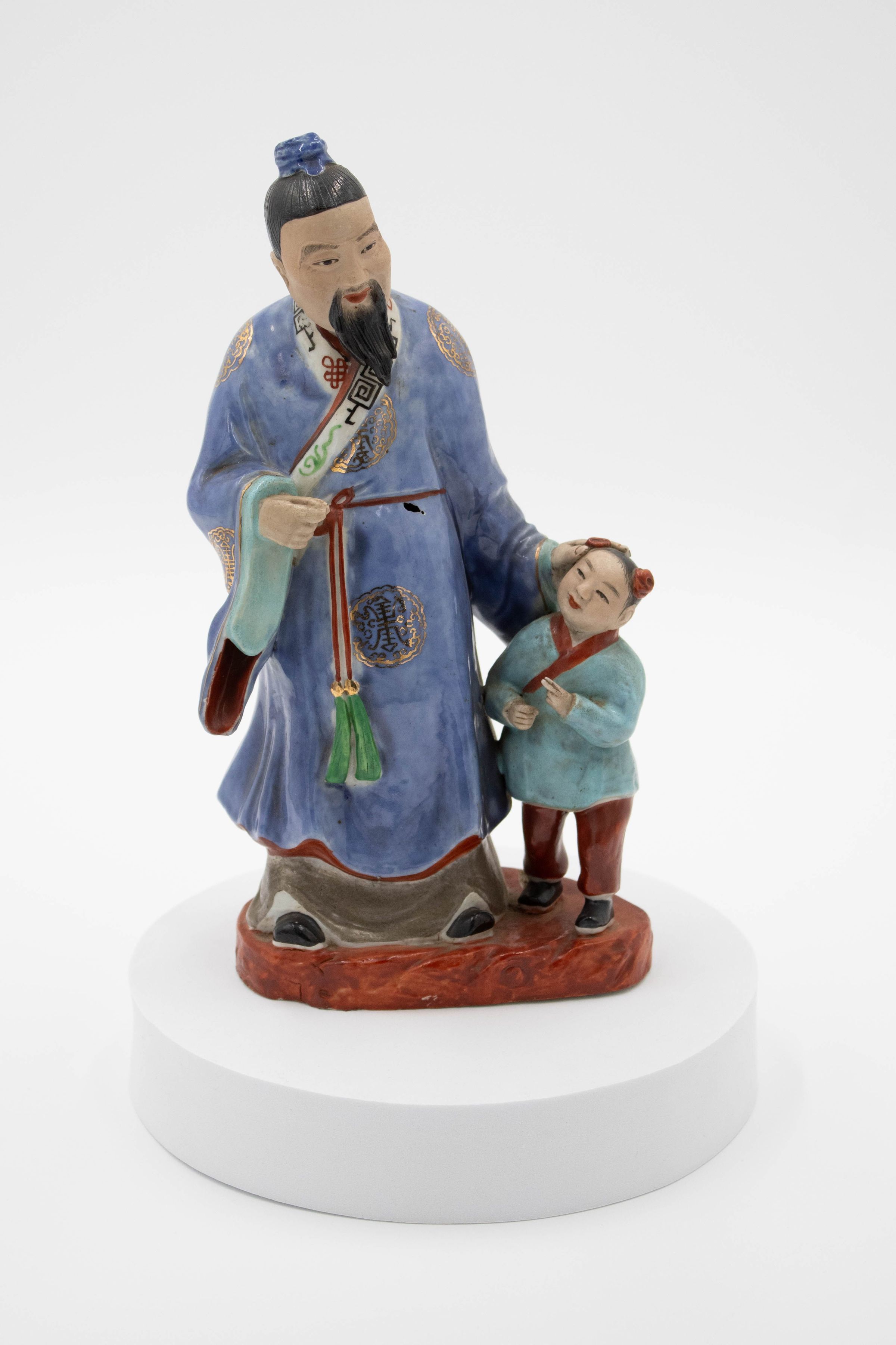 Painted porcelain figurine of Mongolian man and boy.: Image of porcelain figurine of a Mongolian man dressed in a blue robe with gold foil Mongolian symbols on it. He stands above a little boy who is wearing a light blue top with red details and red pants. The man's hair is in a bun atop his head with a blue covering and the boy has two buns both with red coverings. They are standing on a red base.