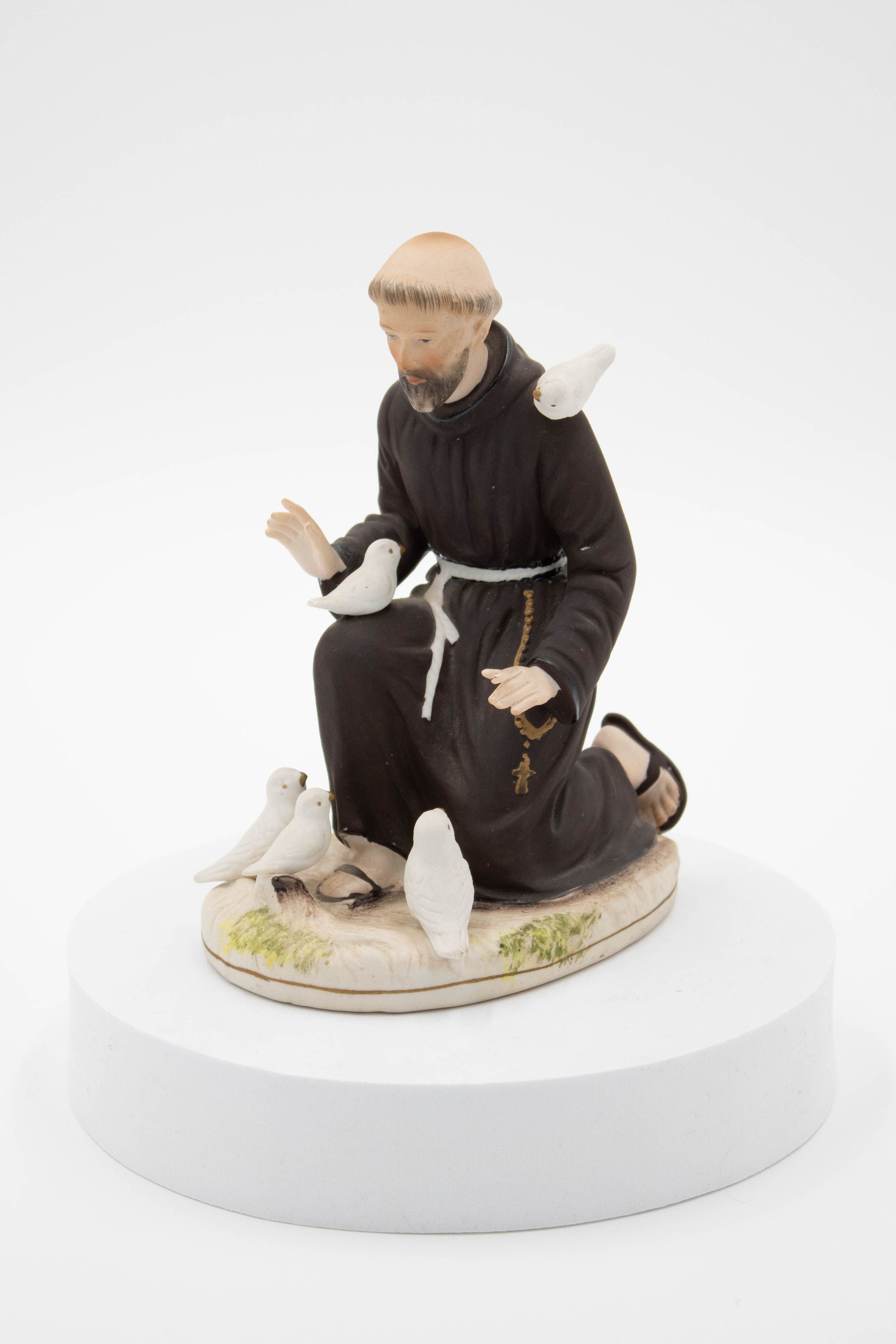 Painted Porcelain Figurine of St. Francis of Assisi: Image of the figurine. The base is porcelain and is of a patchy area of ground, largely being white/brown with small patches of grass. The porcelain figurine depicts Saint Francis of Assisi as a fair-skinned bearded Catholic monk with a tonsure kneeling and preaching to three white birds. Another white bird is on his bent knee, while another is on his left shoulder. All the birds are looking up at him as he speaks. He wears sandals and has a white cloth belt around his habit, as well as a large rosary at his hip.
